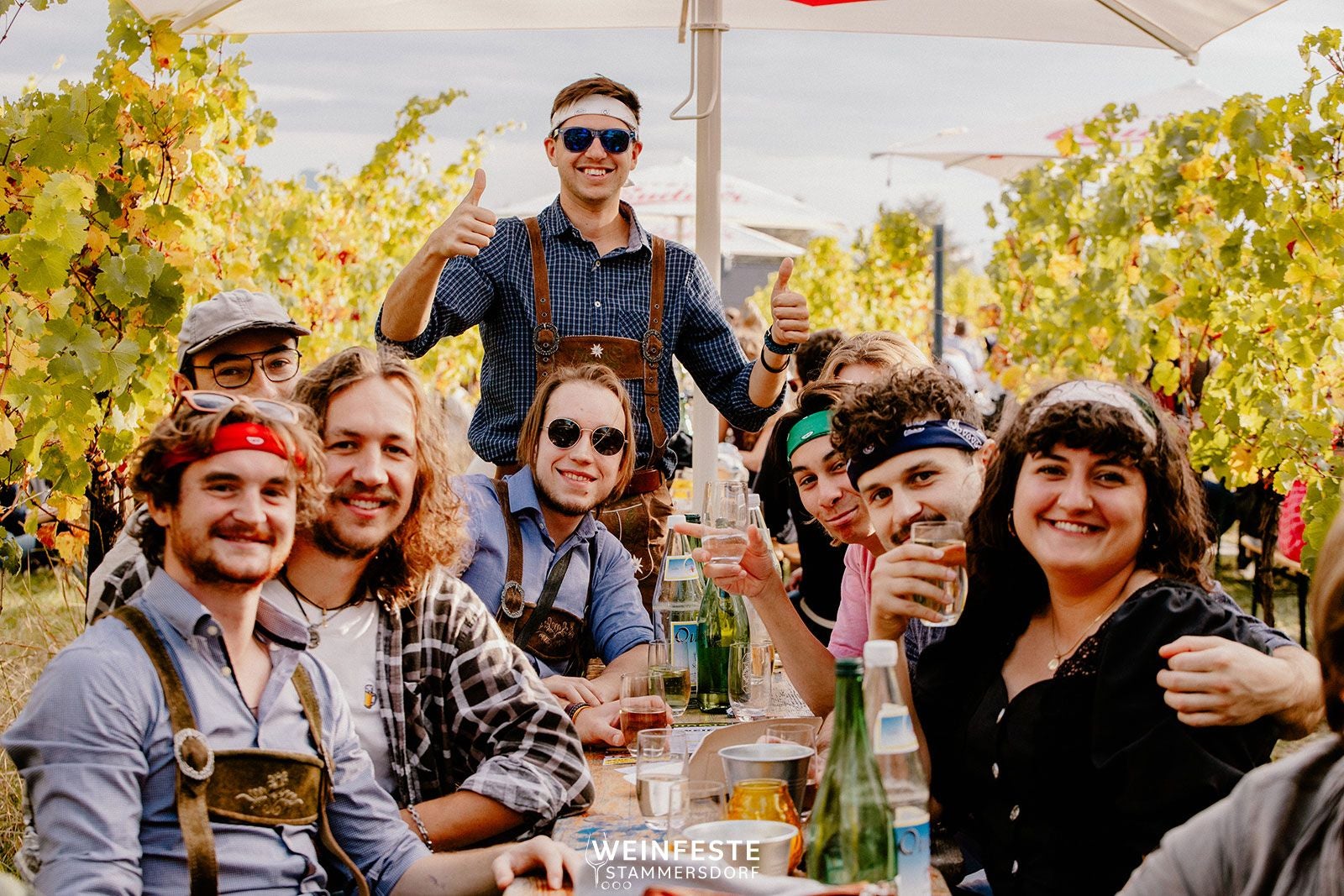 Junge Menschen feiern ausgelassen an einem langen Tisch im Weingarten, trinken und lachen.