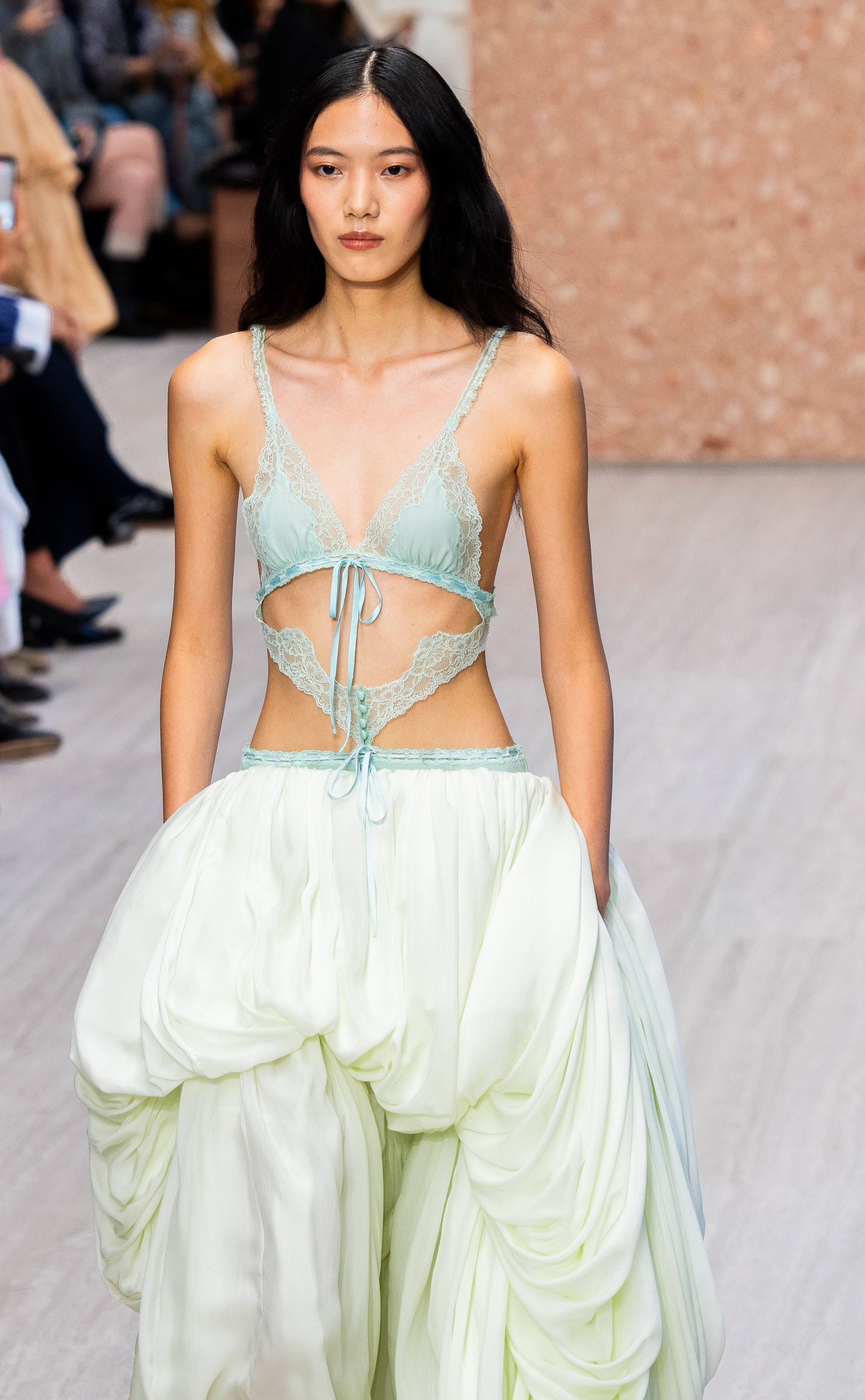 Model in mintgrünem Spitzenoberteil und weiter, geraffter, heller Hose auf dem Laufsteg.