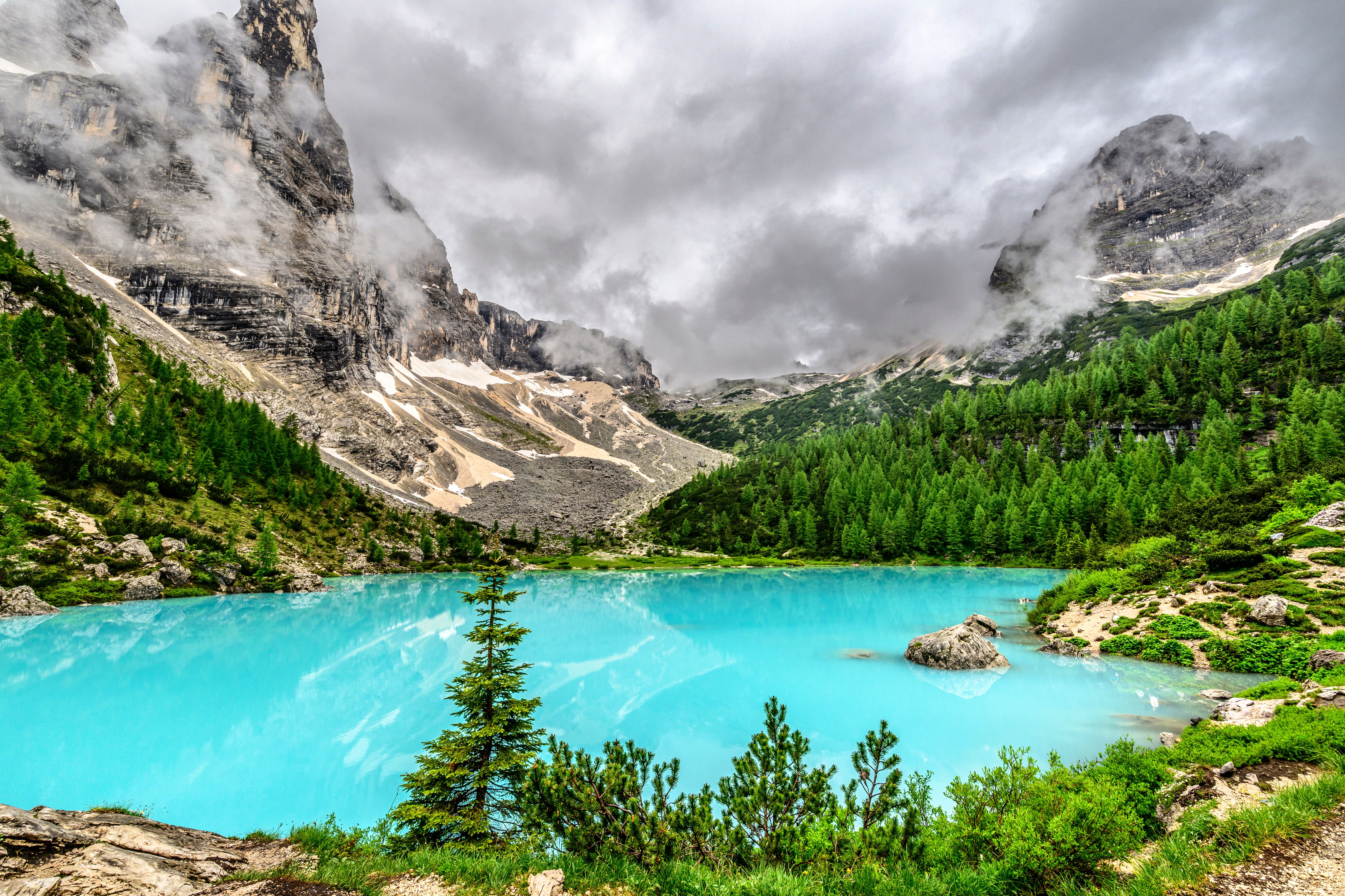 Der türkisfarbene Lago di Sorapis liegt vor bewaldeten Berghängen und wolkenverhangenen Dolomitengipfeln.