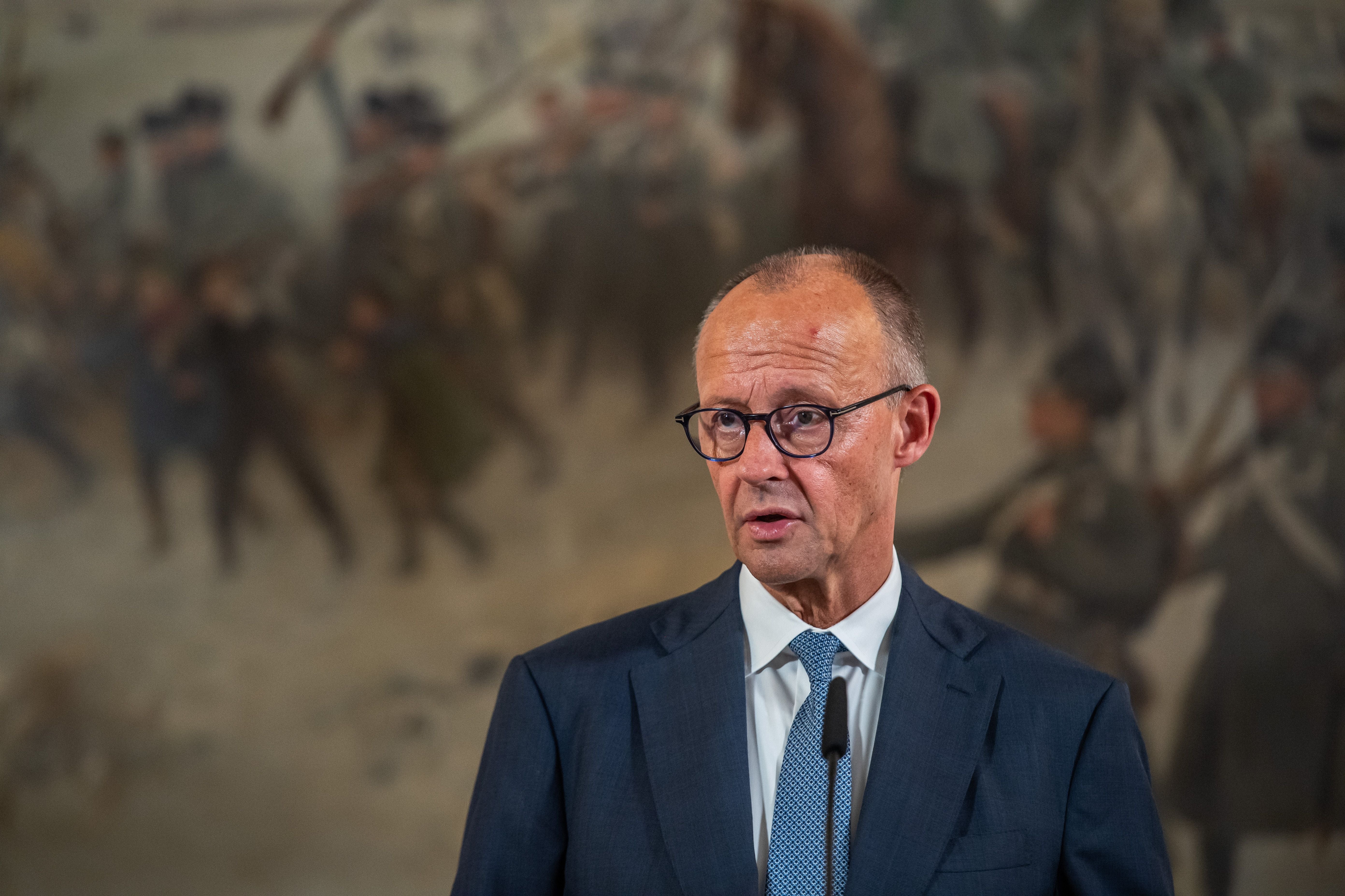 Ein Mann im Anzug spricht vor Mikrofon, im Hintergrund unscharfes Wandgemälde mit Soldaten.