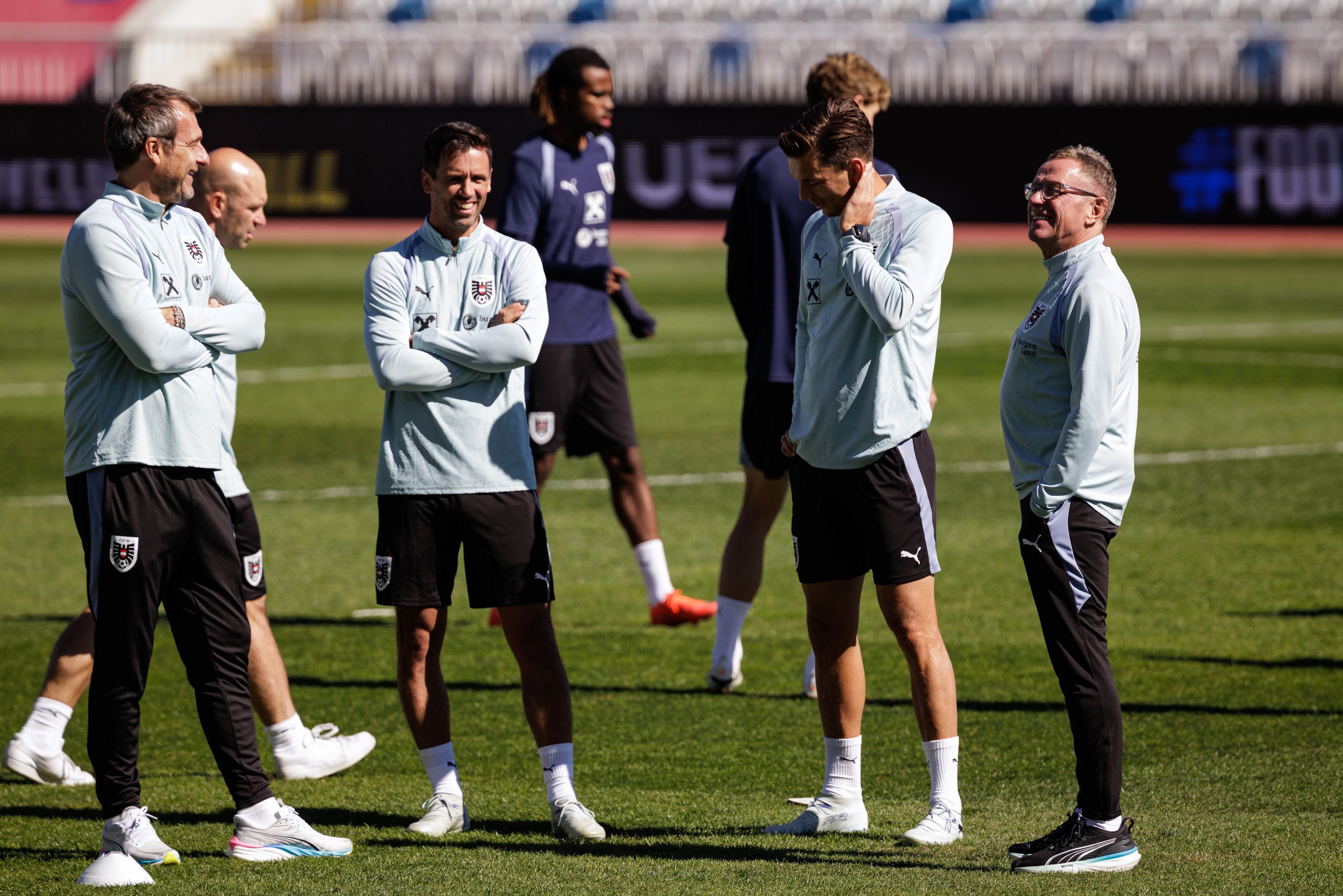 Vier österreichische Fußballtrainer stehen lachend auf einem Trainingsplatz im Sonnenschein.