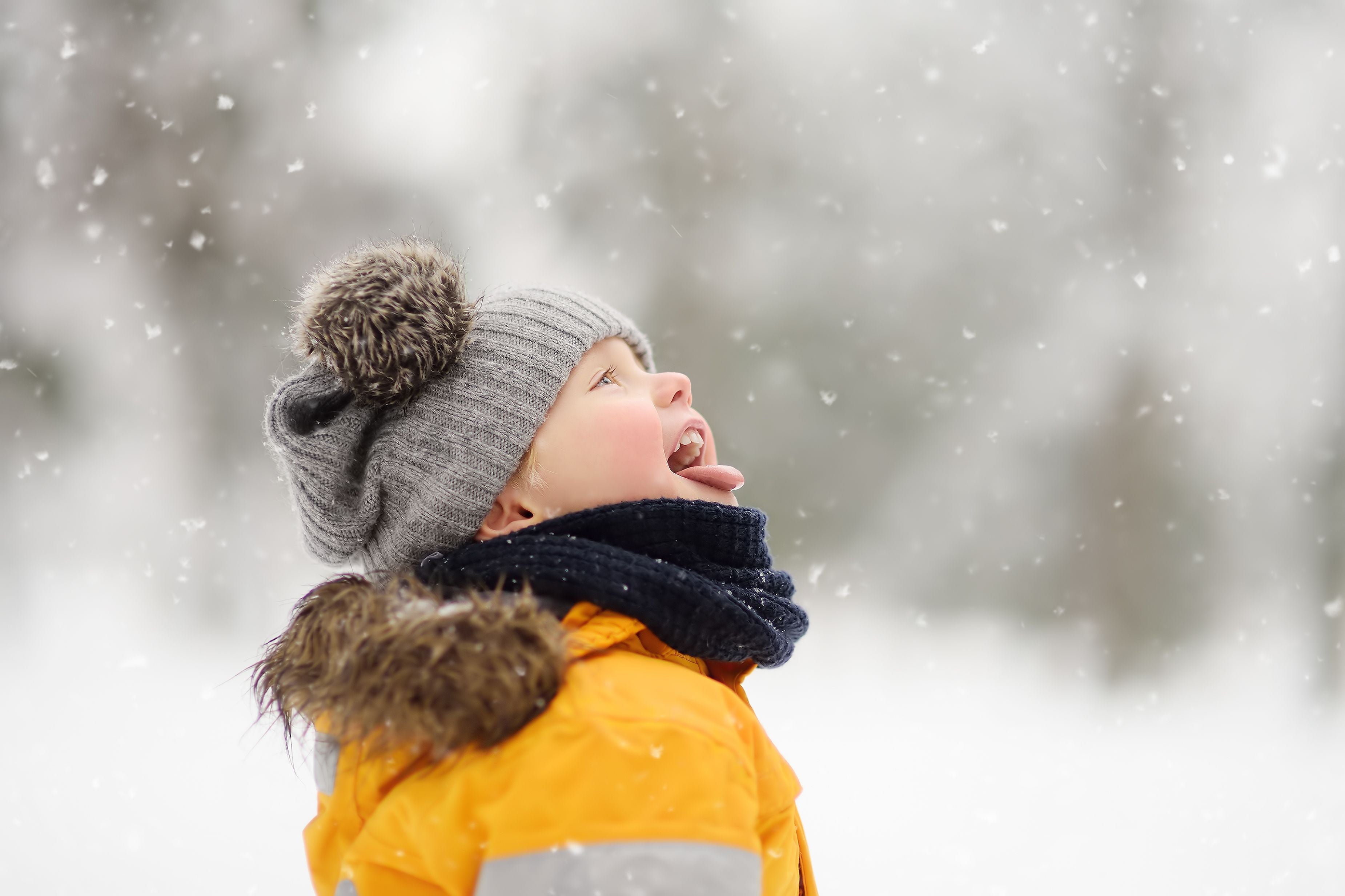 Kind zieht im Wintermantel und Mütze die Zunge raus und fängt fallende Schneeflocken.