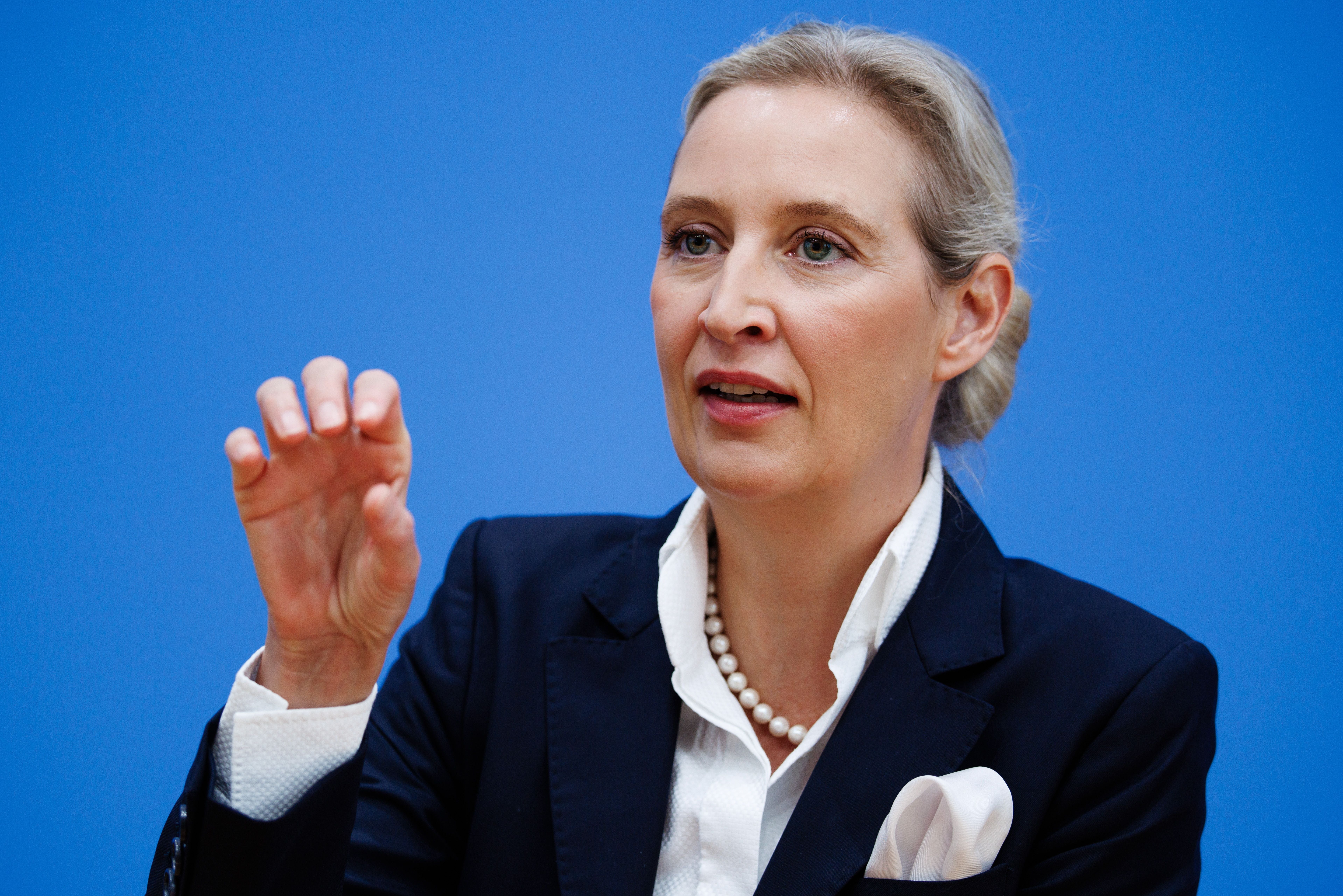 Frau im Anzug spricht gestikulierend vor blauem Hintergrund bei einer Pressekonferenz.