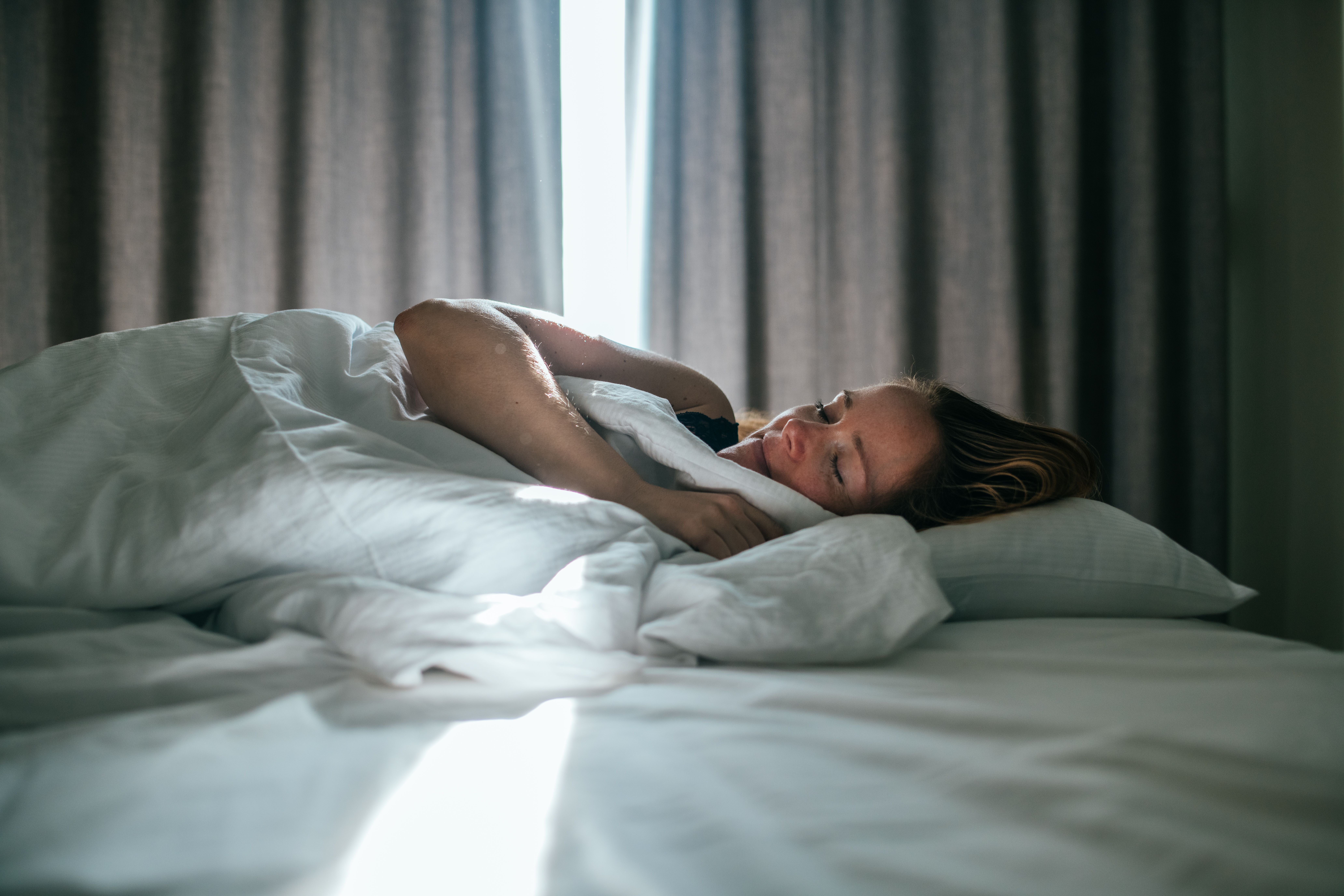 Frau schläft entspannt im Bett, Sonnenlicht fällt durch die Vorhänge im Hintergrund.