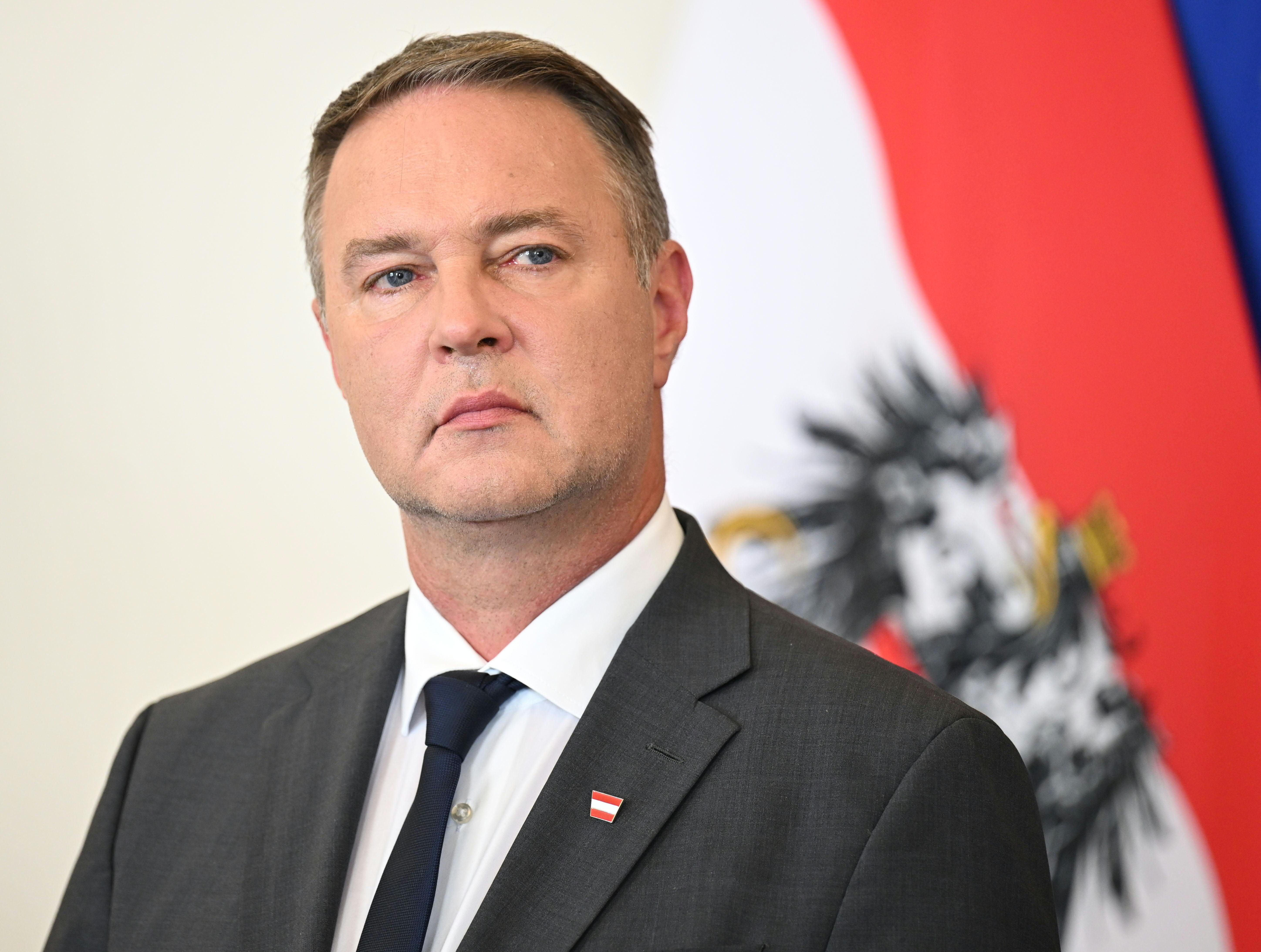 Ein Mann im Anzug steht vor einer österreichischen Flagge und schaut ernst.