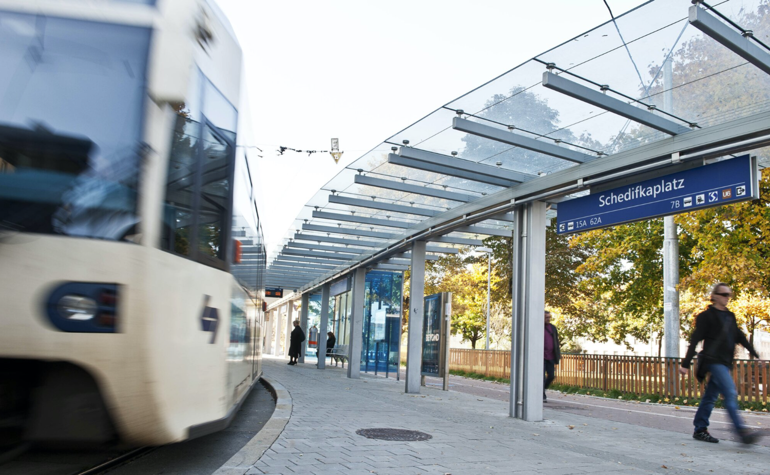 Straßenbahn fährt an der Haltestelle Schedifkaplatz vorbei, Menschen gehen am Bahnsteig entlang.