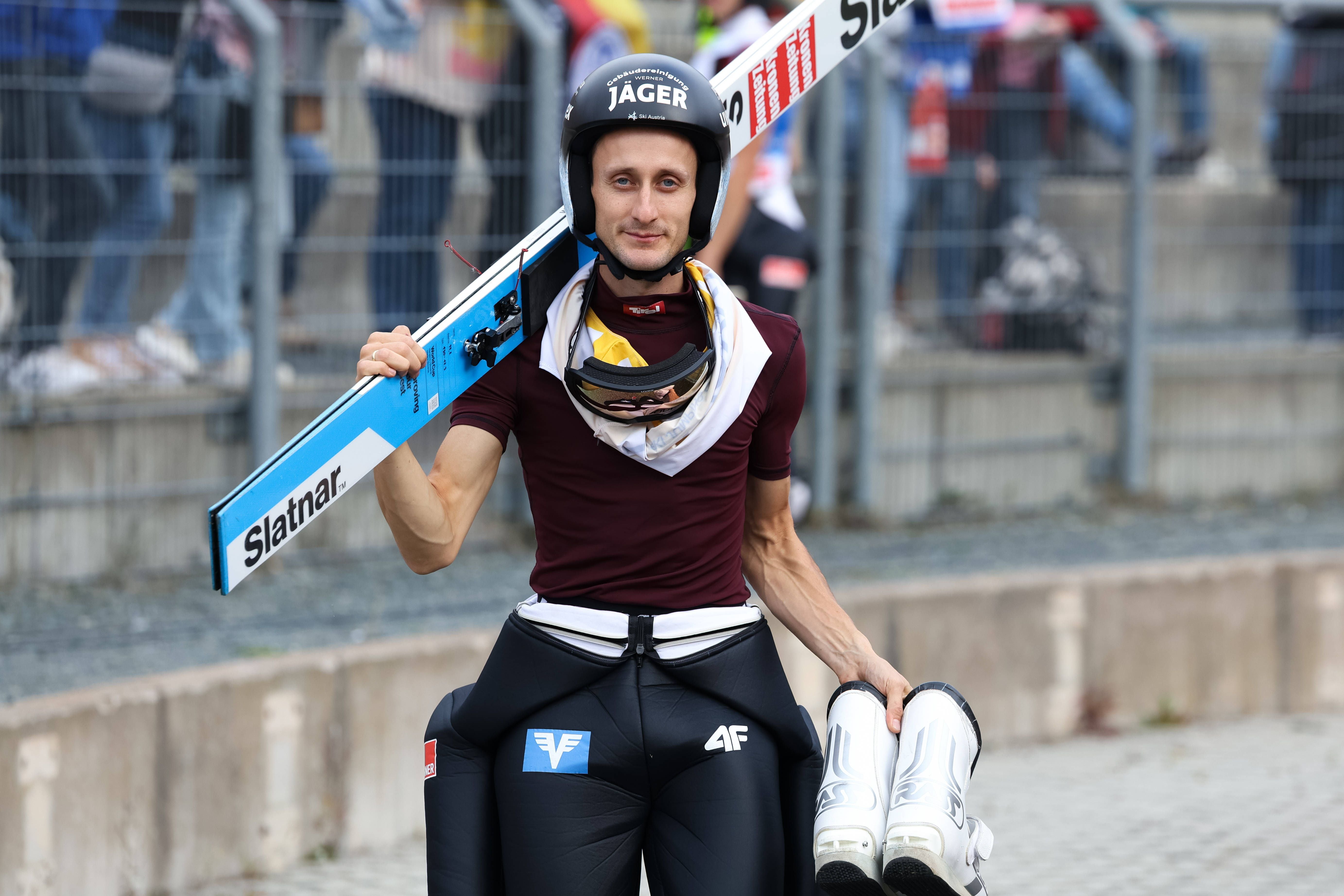 Ein Skispringer mit Helm und Skiern über der Schulter auf dem Weg zur Sprunganlage.