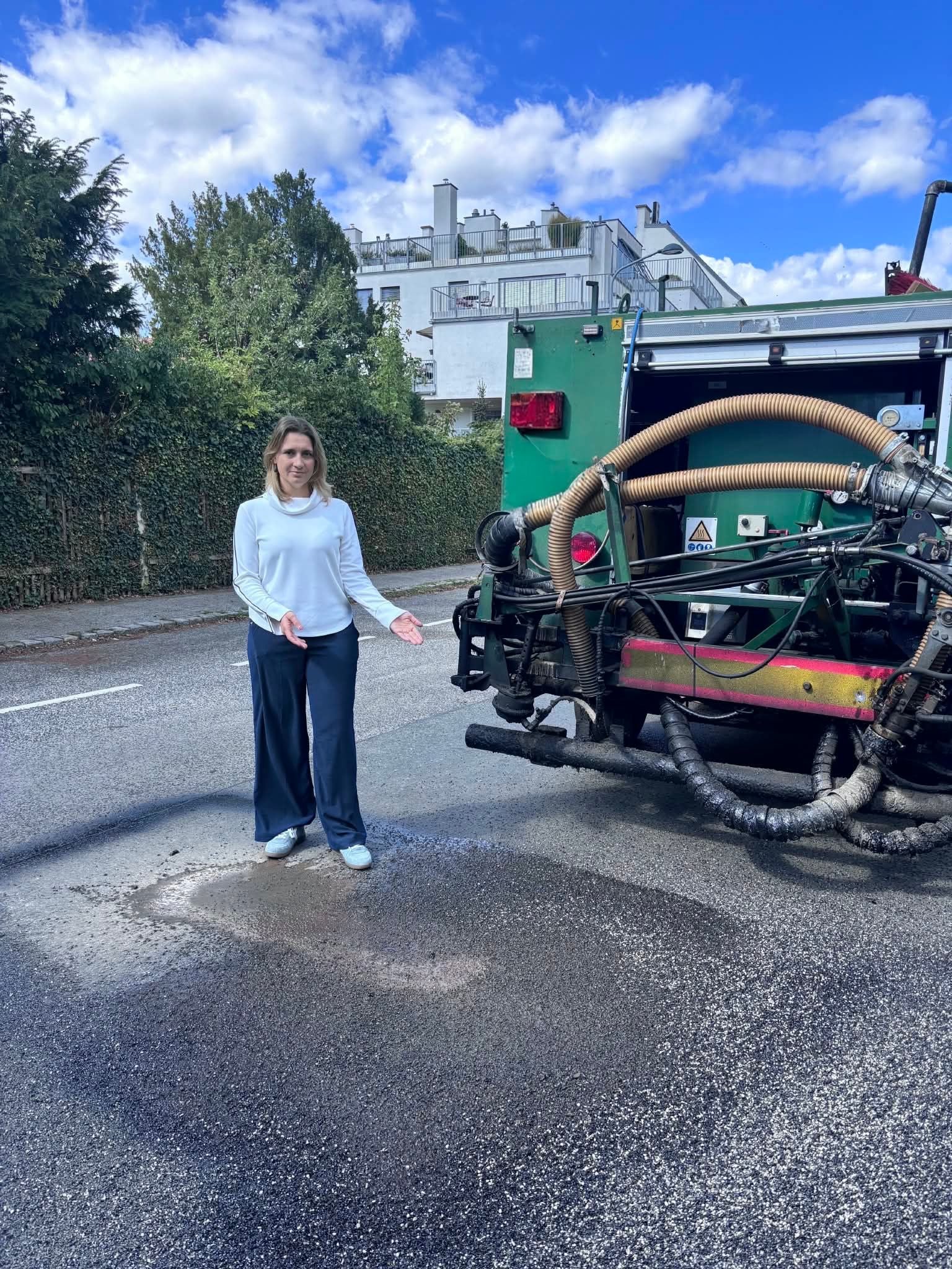Eine Frau steht neben einer Straßenbaumaschine und zeigt auf eine reparierte Asphaltstelle.