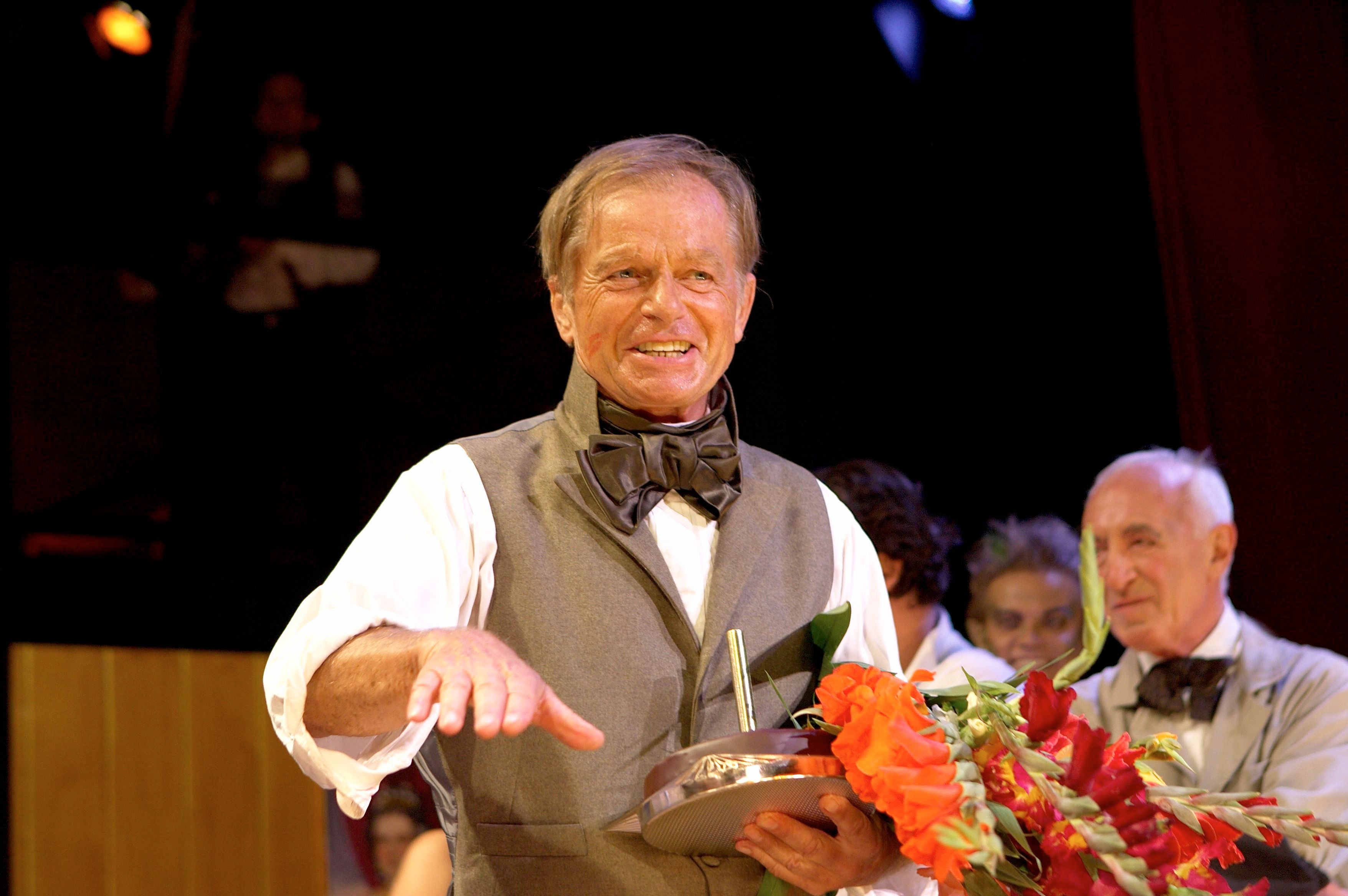 Älterer Mann mit Blumenstrauß und Geschenk lächelt bei einer Feier, mehrere Personen im Hintergrund.