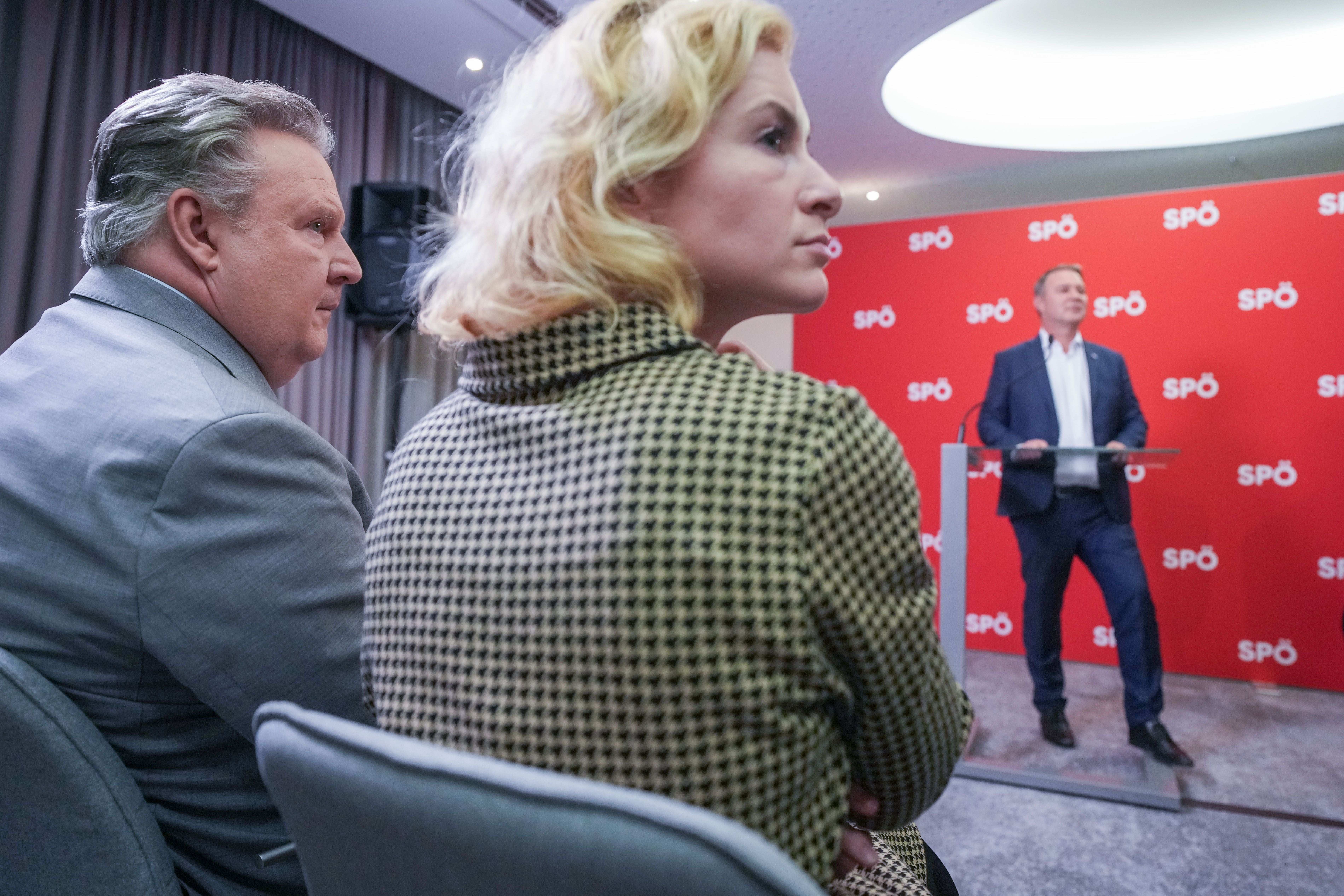 Zwei Personen sitzen, während ein Mann vor roter SPÖ-Wand an einem Rednerpult steht.