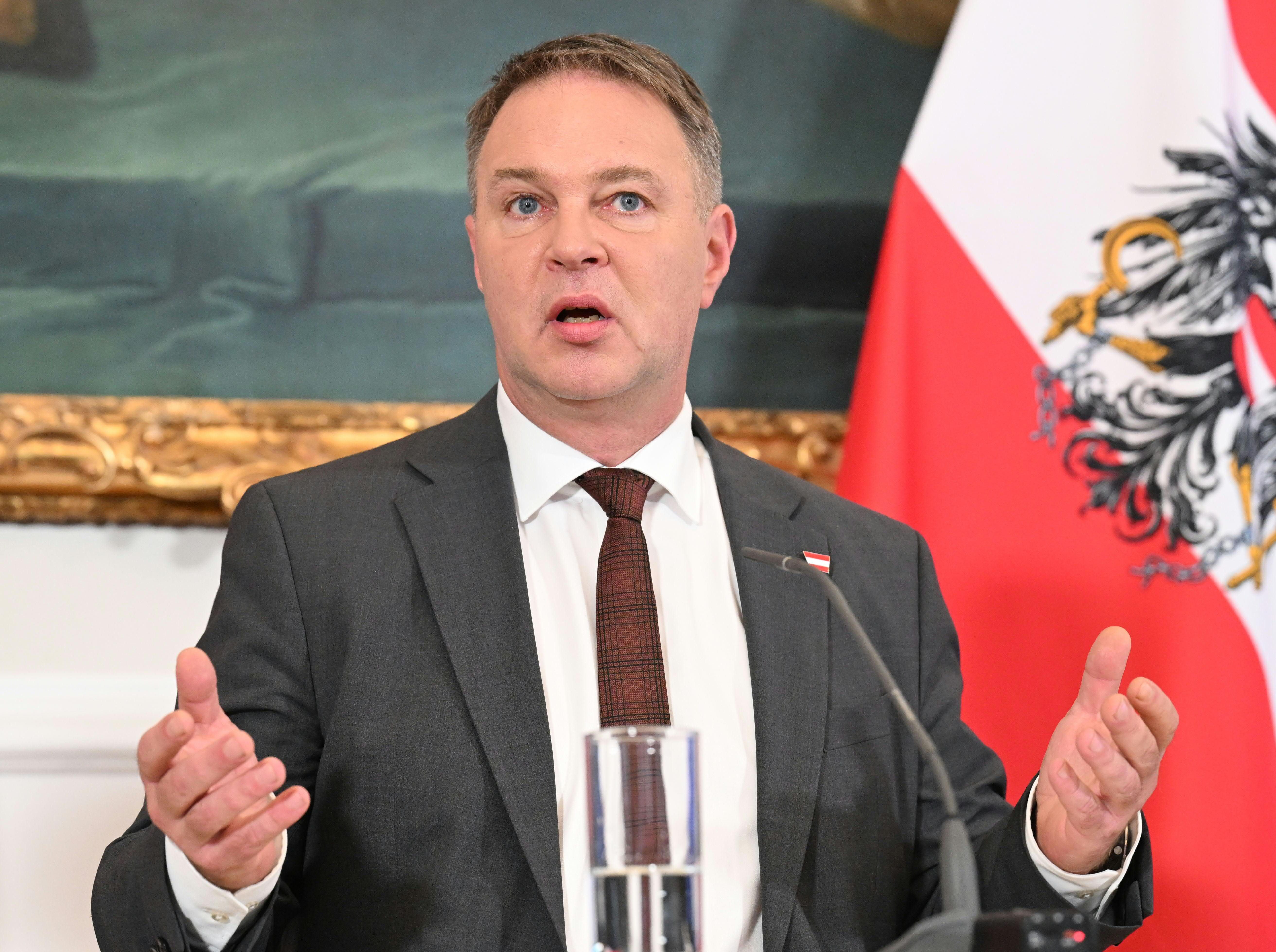 Ein Mann im Anzug spricht bei einer Pressekonferenz, im Hintergrund eine österreichische Flagge.