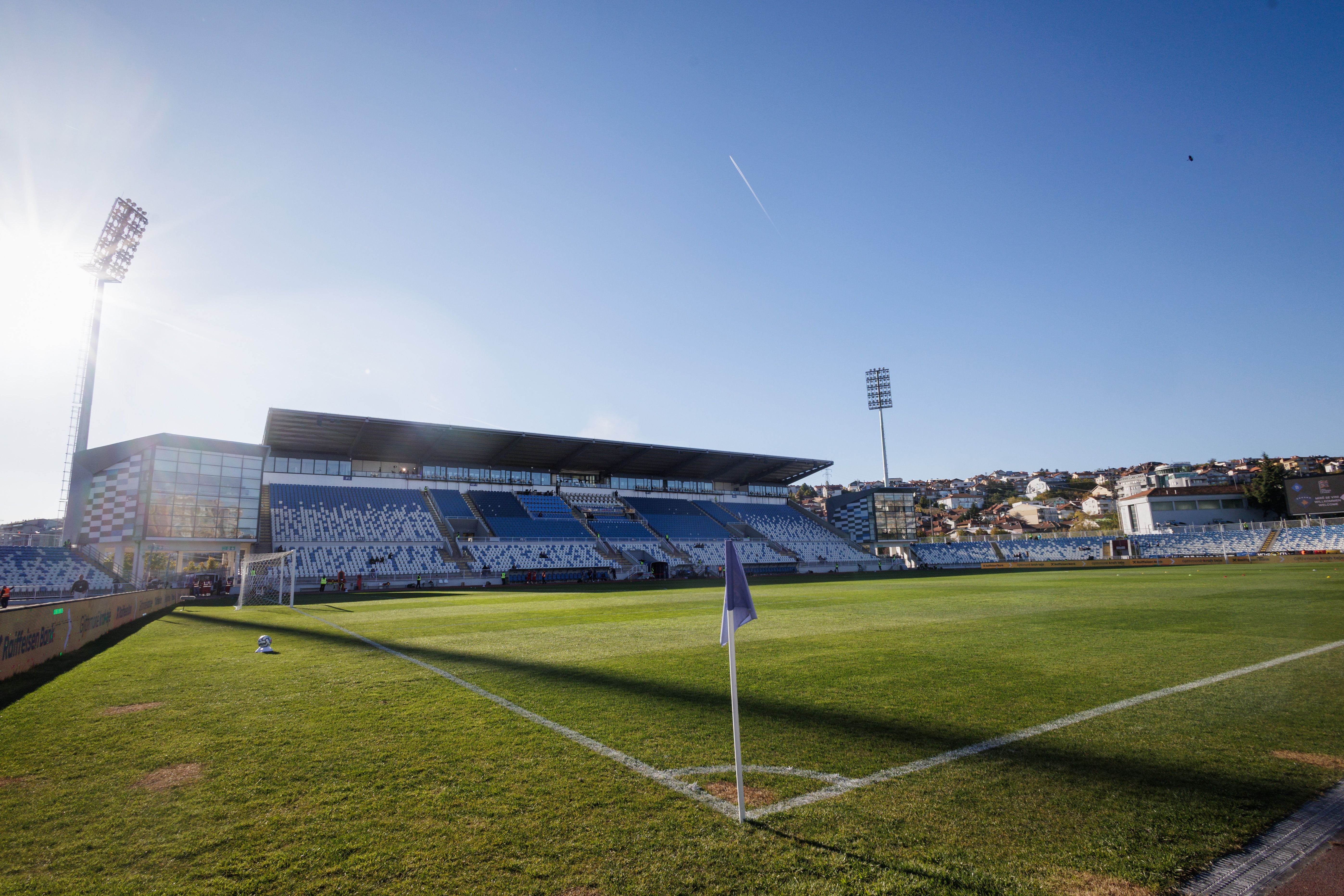 Blick von der Eckfahne über das grüne Spielfeld des Fadil-Vokrri-Stadions in Pristina unter blauem Sonnenschein.