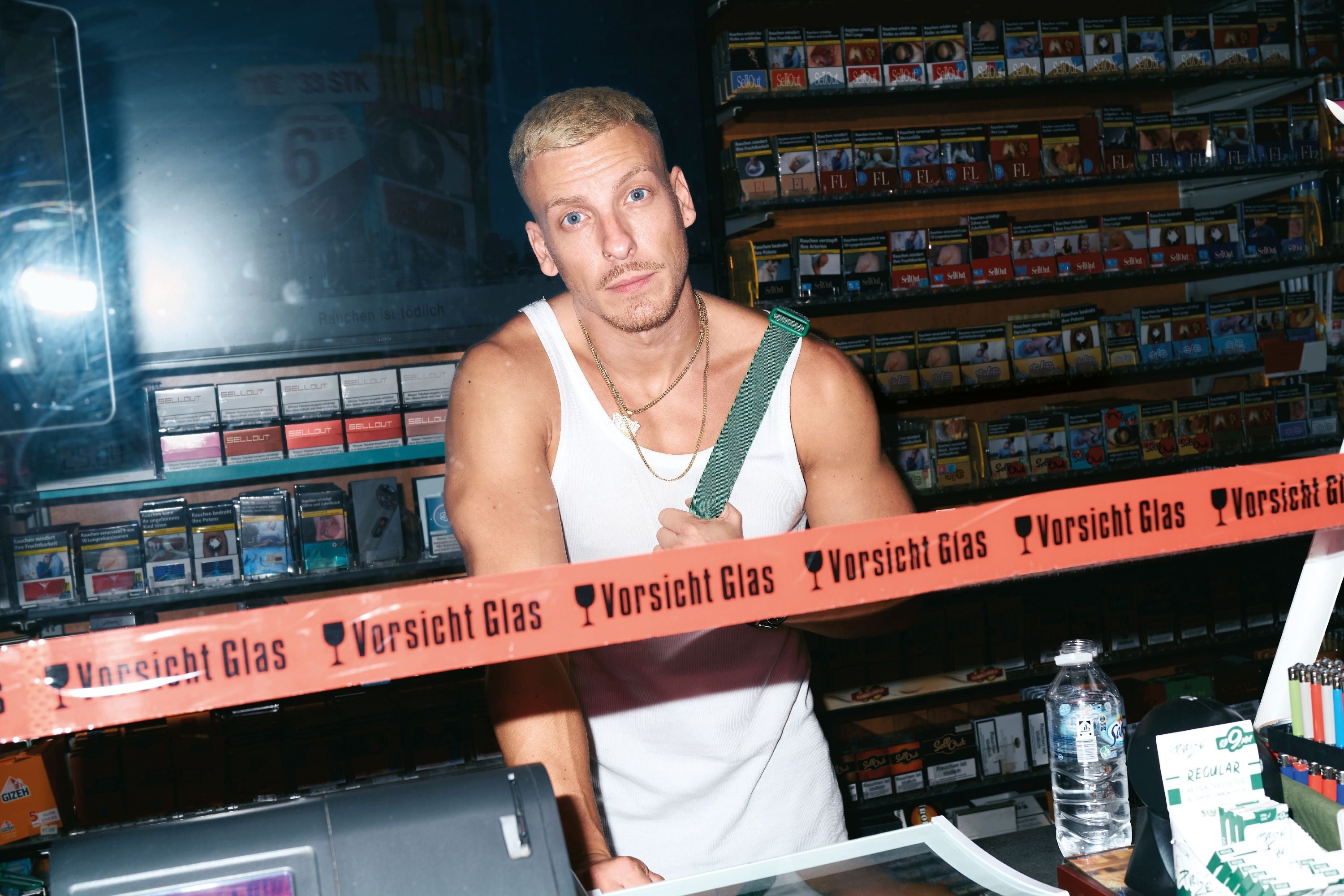 Mann im weißen Tanktop steht hinter einer Theke mit Warnband "Vorsicht Glas".