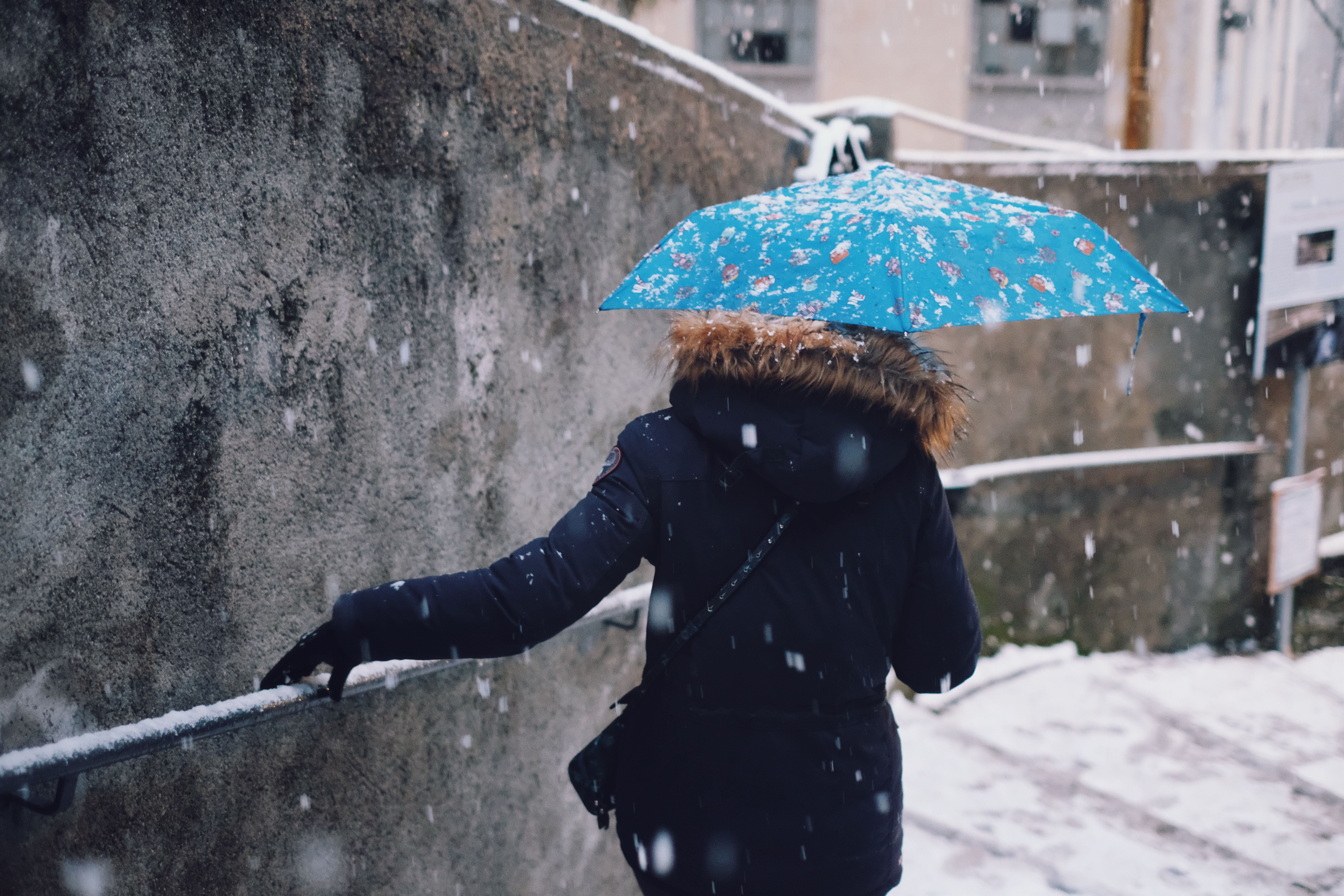 Mädchen mit blauem Regenschirm läuft bei Schneefall eine Treppe im Dorf hinunter.