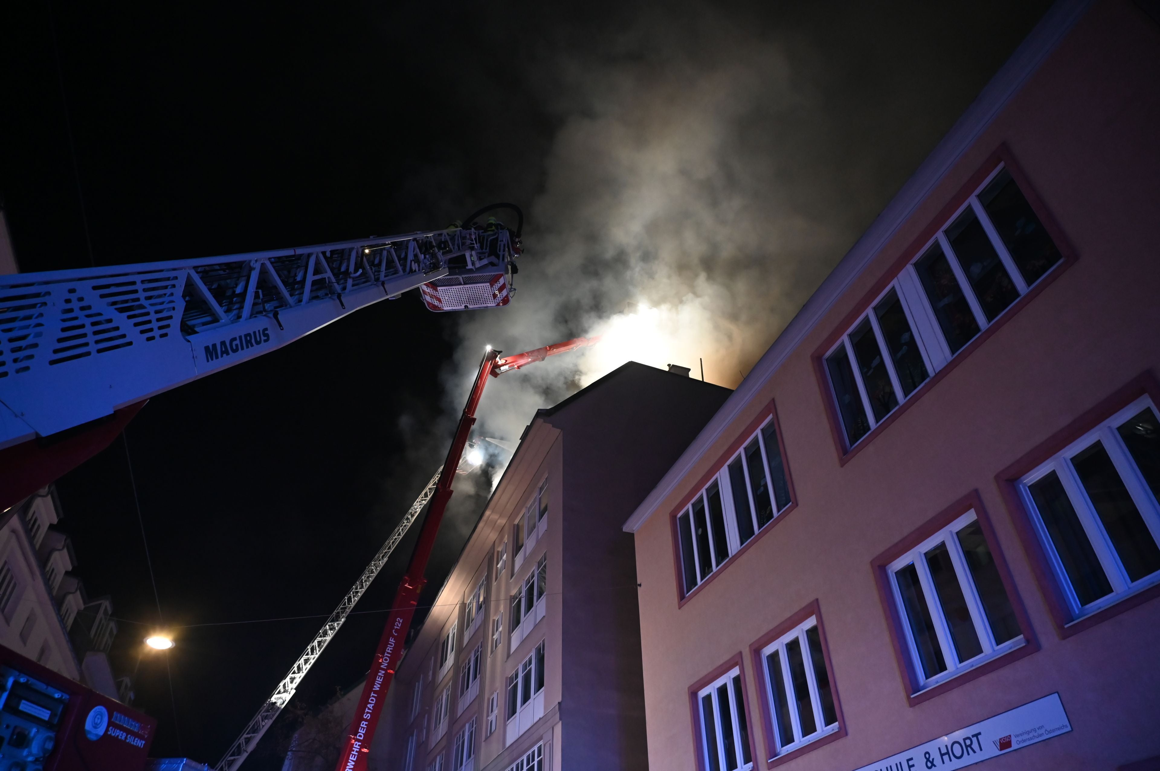 Nachtaufnahme eines brennenden Gebäudes mit Feuerwehrleitern und starkem Rauch am Himmel.