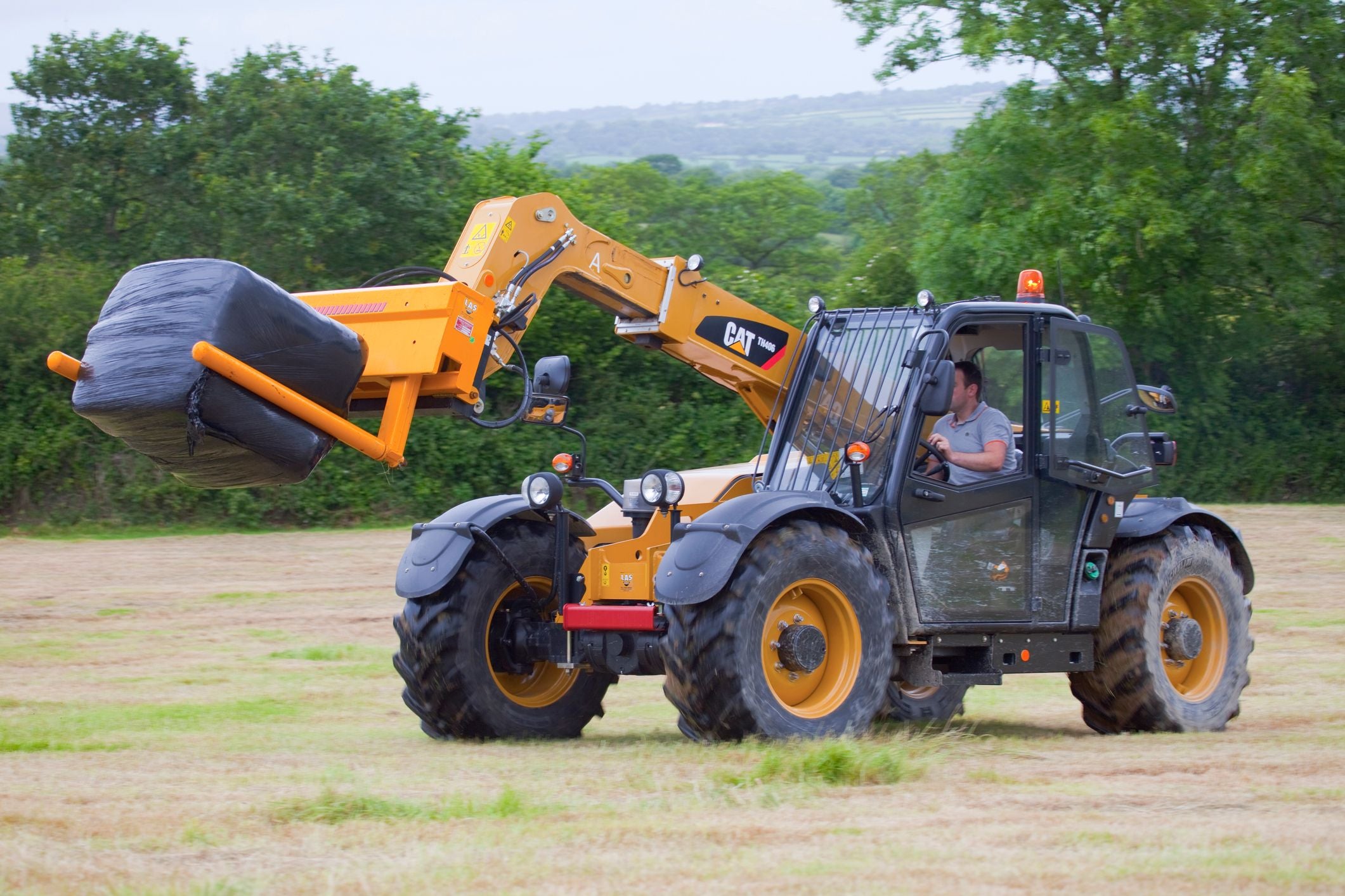 Ein CAT Teleskoplader transportiert einen in Folie gewickelten Heuballen über ein Feld.