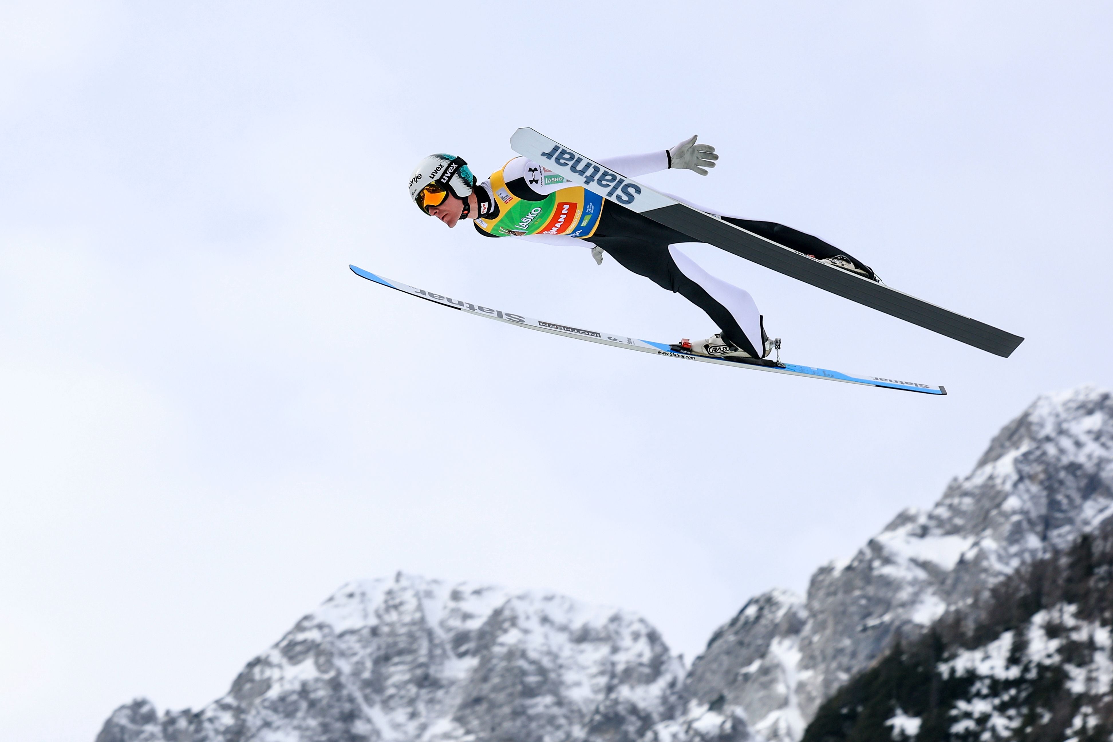 Skispringer fliegt durch die Luft vor schneebedeckten Bergen und bewölktem Himmel.