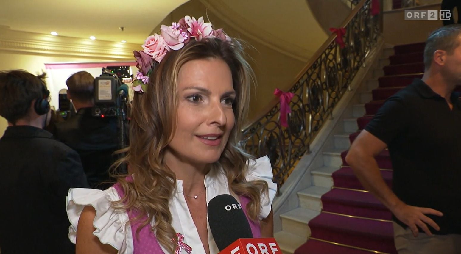 Frau mit Blumenkranz im Haar gibt ein Interview auf einer Treppe mit rotem Teppich.