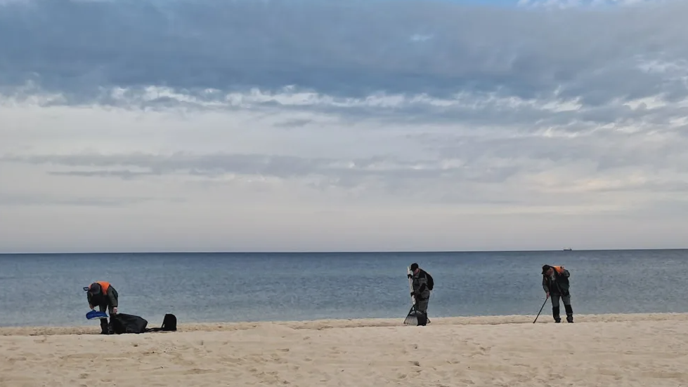 Drei Personen reinigen einen Strand vor der Ostsee, jeweils mit Ausrüstung und Müllsäcken.