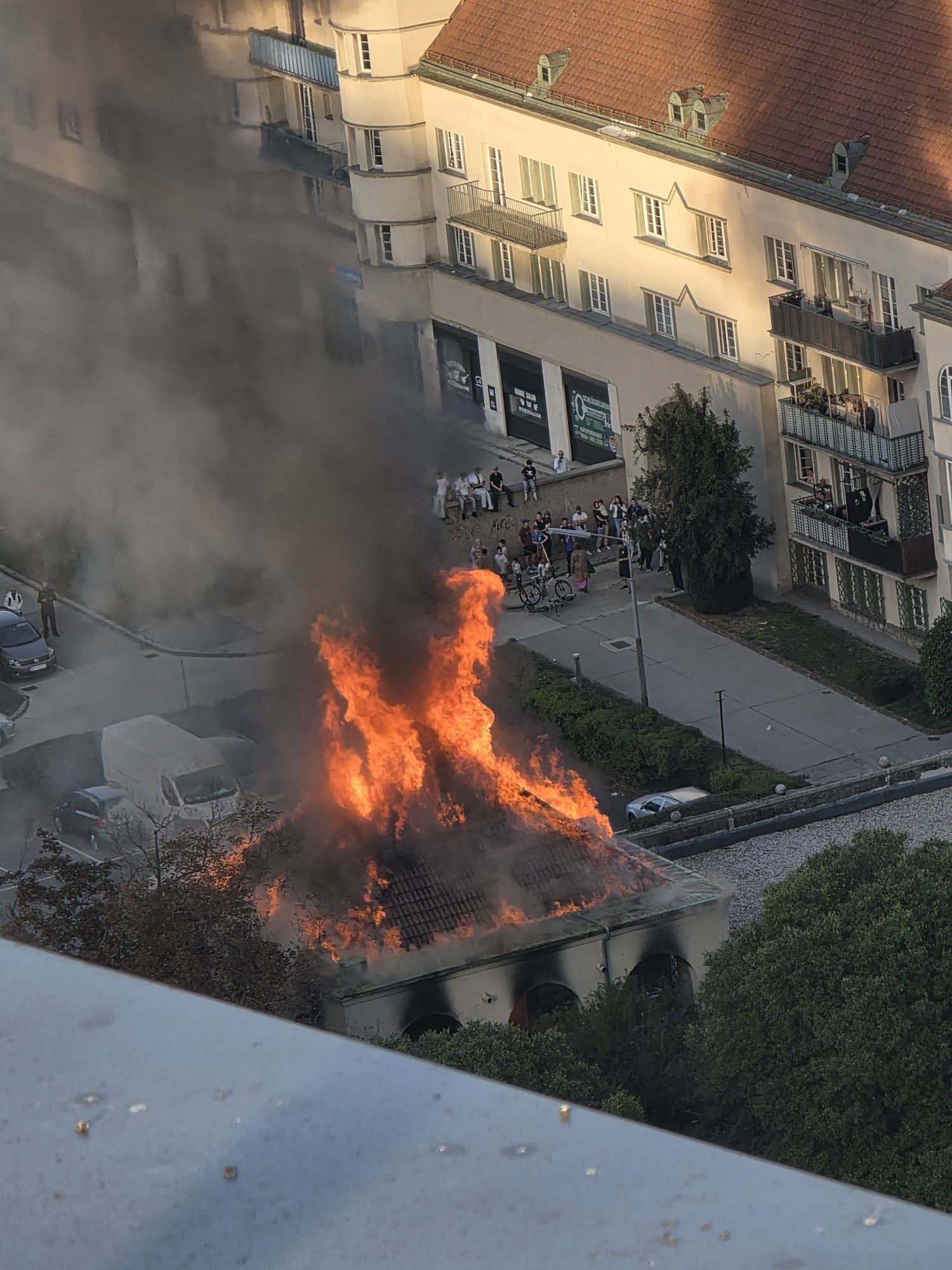Stark brennendes Gebäude mit hoher Rauchentwicklung, Menschen beobachten das Feuer aus sicherer Entfernung.