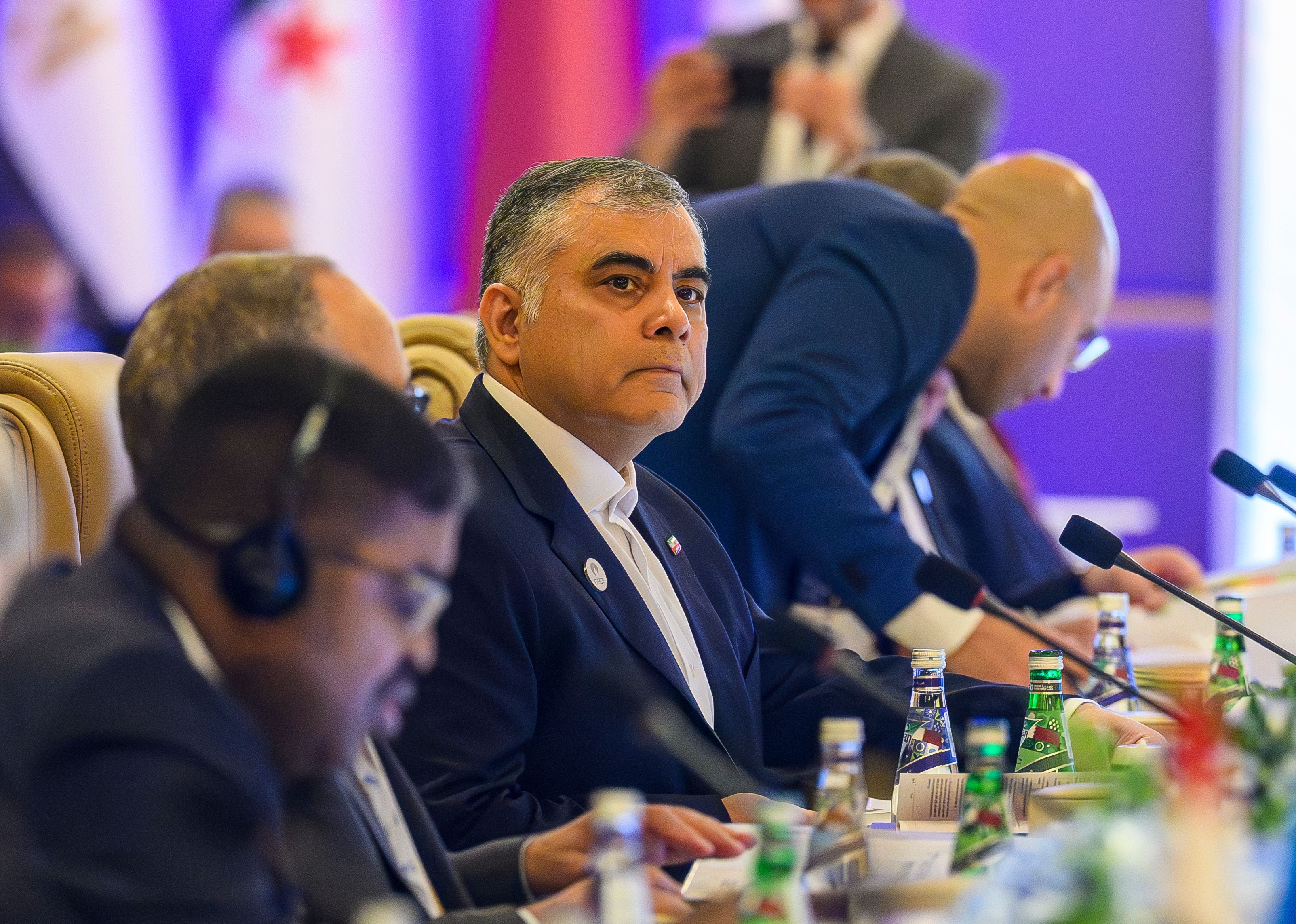 Mehrere Männer sitzen bei einer internationalen Konferenz an einem Tisch mit Mikrofonen und Wasserflaschen.