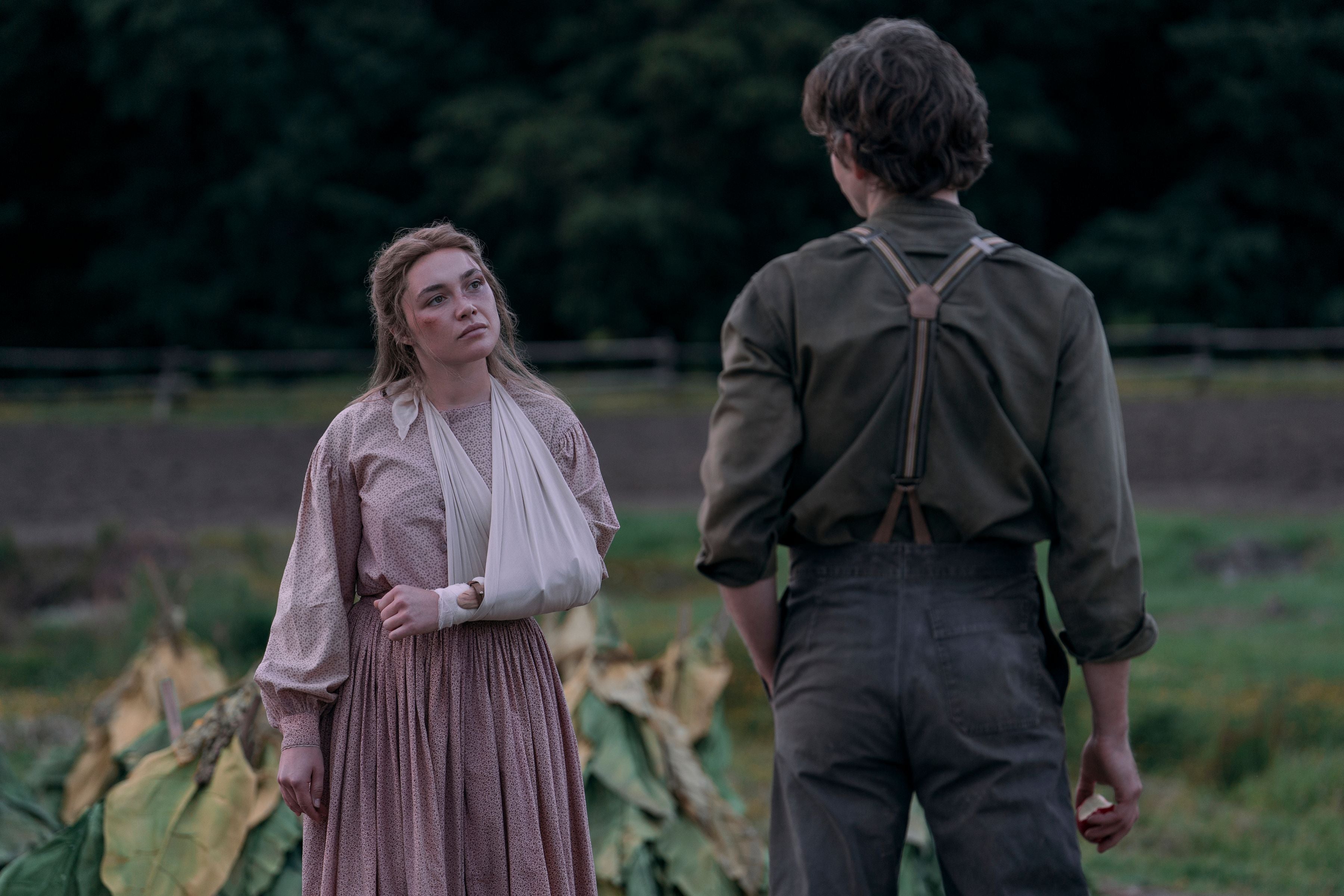 Eine Frau mit Armschlinge steht einem Mann auf einem Feld gegenüber, im Hintergrund Bäume.