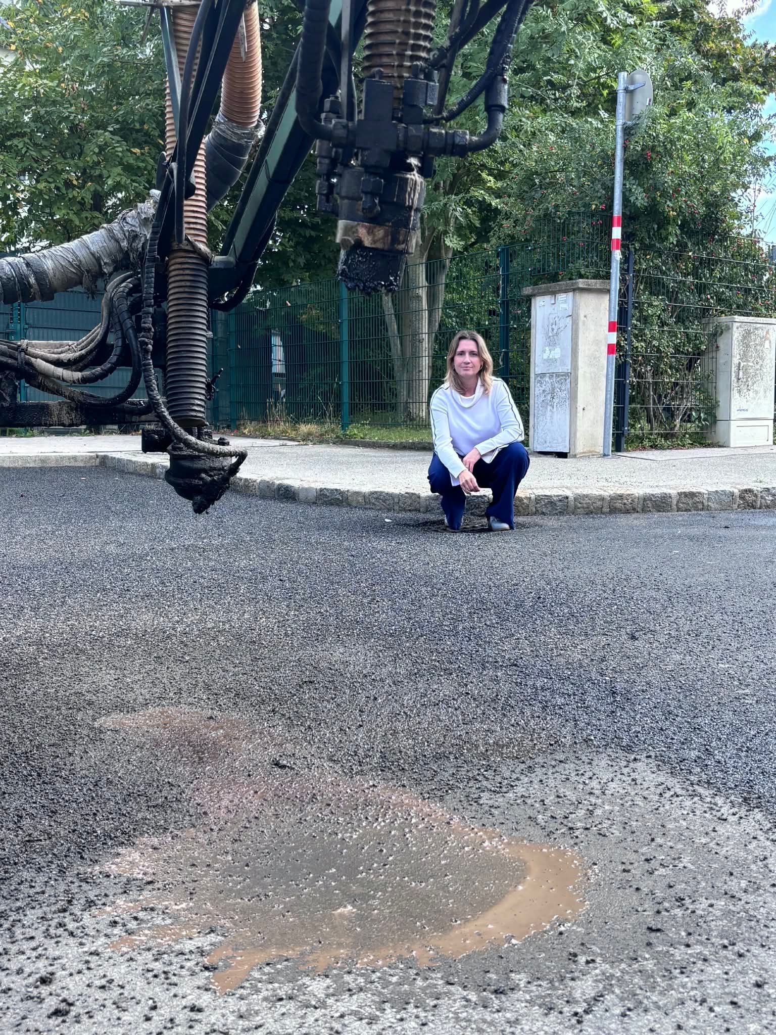 Eine Frau hockt auf Gehweg neben einer Asphaltiermaschine und frischem Straßenbelag.