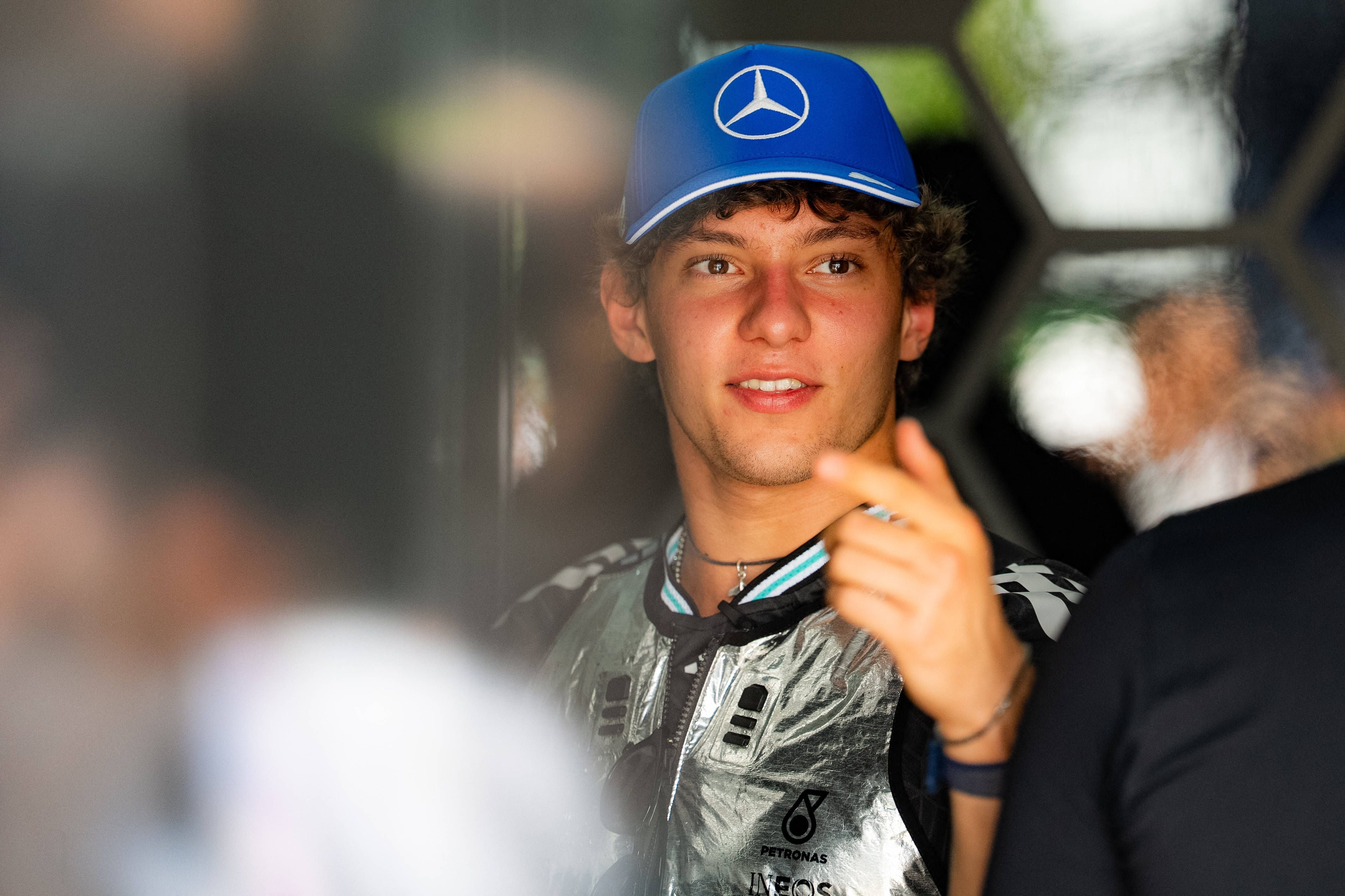 Ein junger Mann mit blauer Mercedes-Mütze und silberner Jacke zeigt mit dem Finger und lächelt.