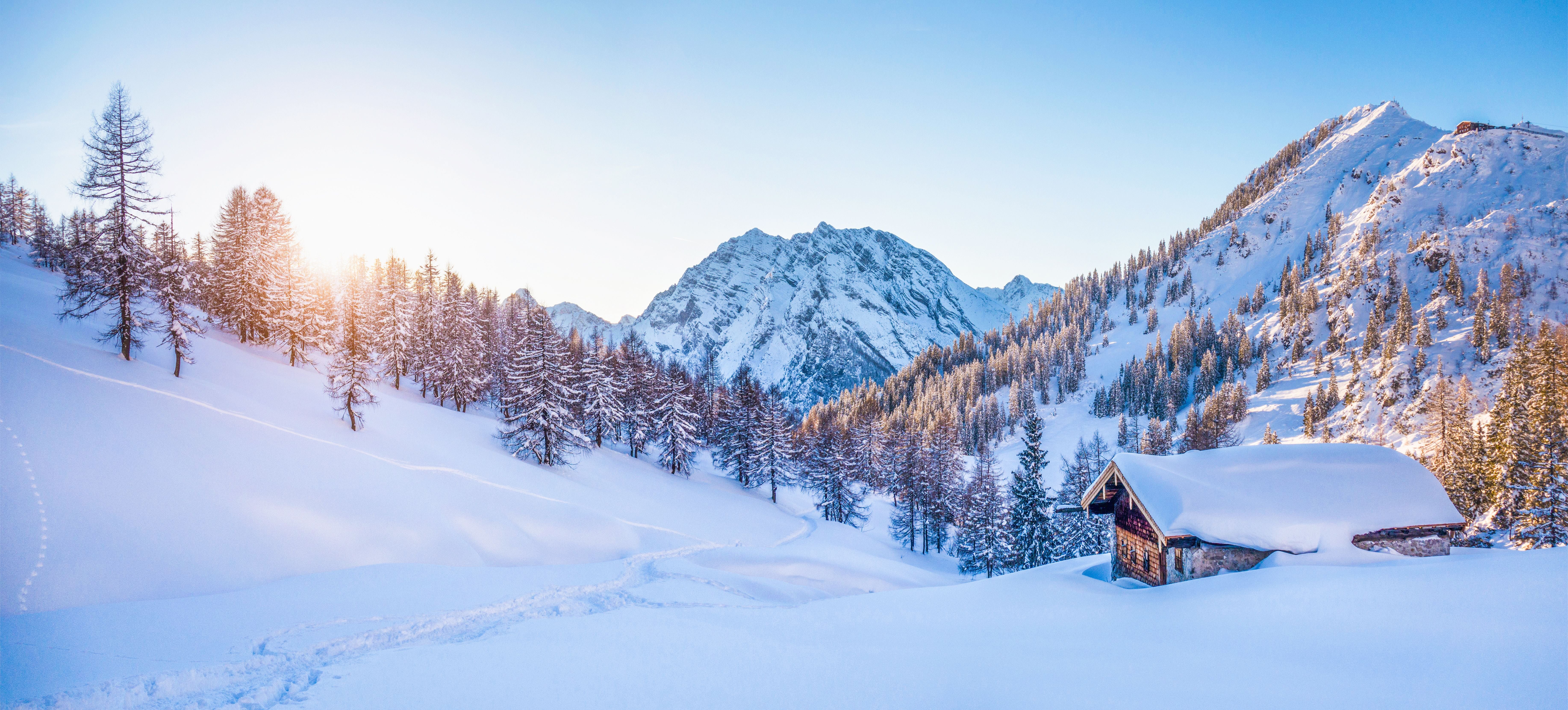 Verschneite Berghütte und Bäume im Sonnenuntergang in winterlicher Alpenlandschaft.