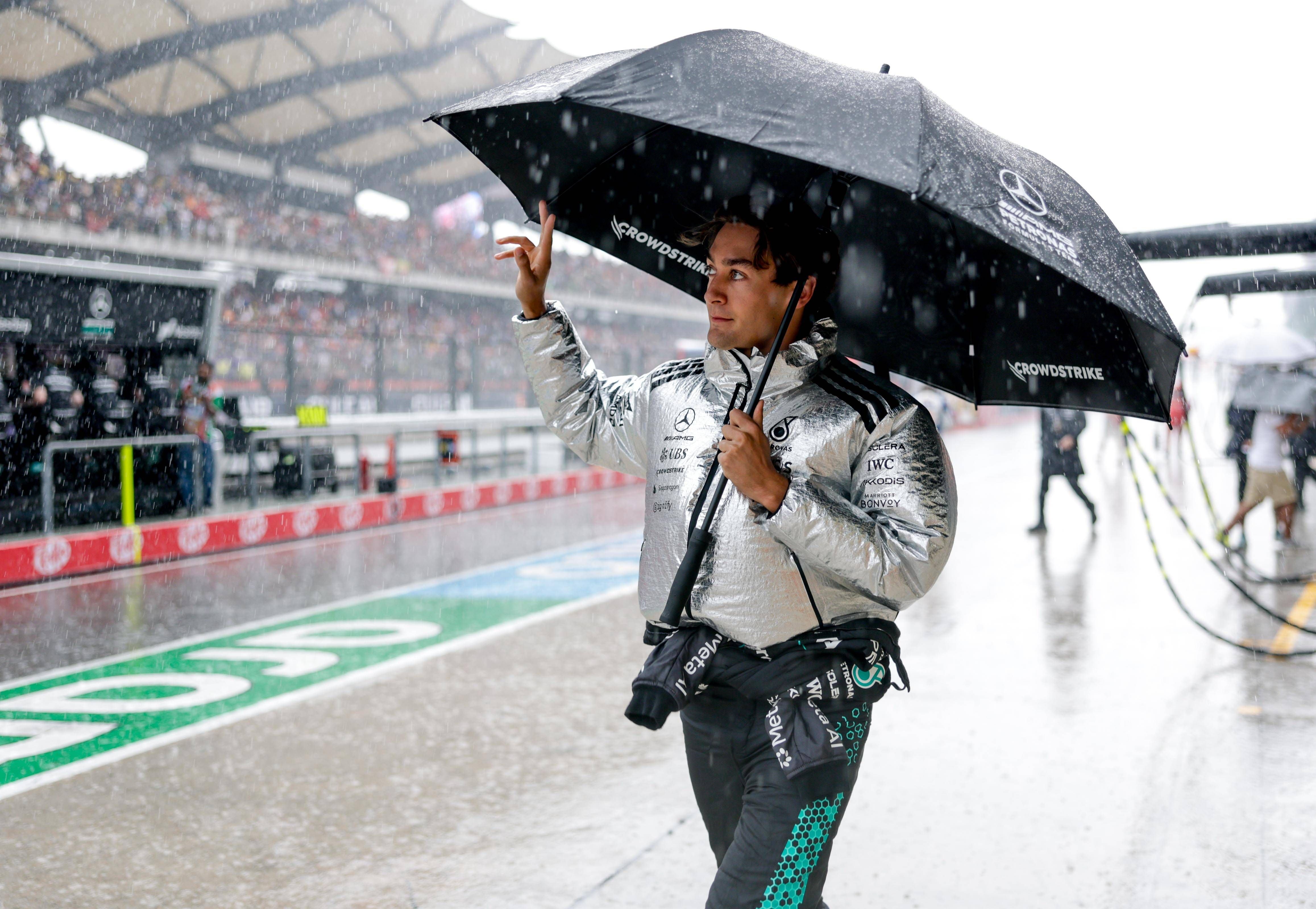 Ein Mann mit silberner Jacke hält einen schwarzen Regenschirm im Regen auf einer Rennstrecke.