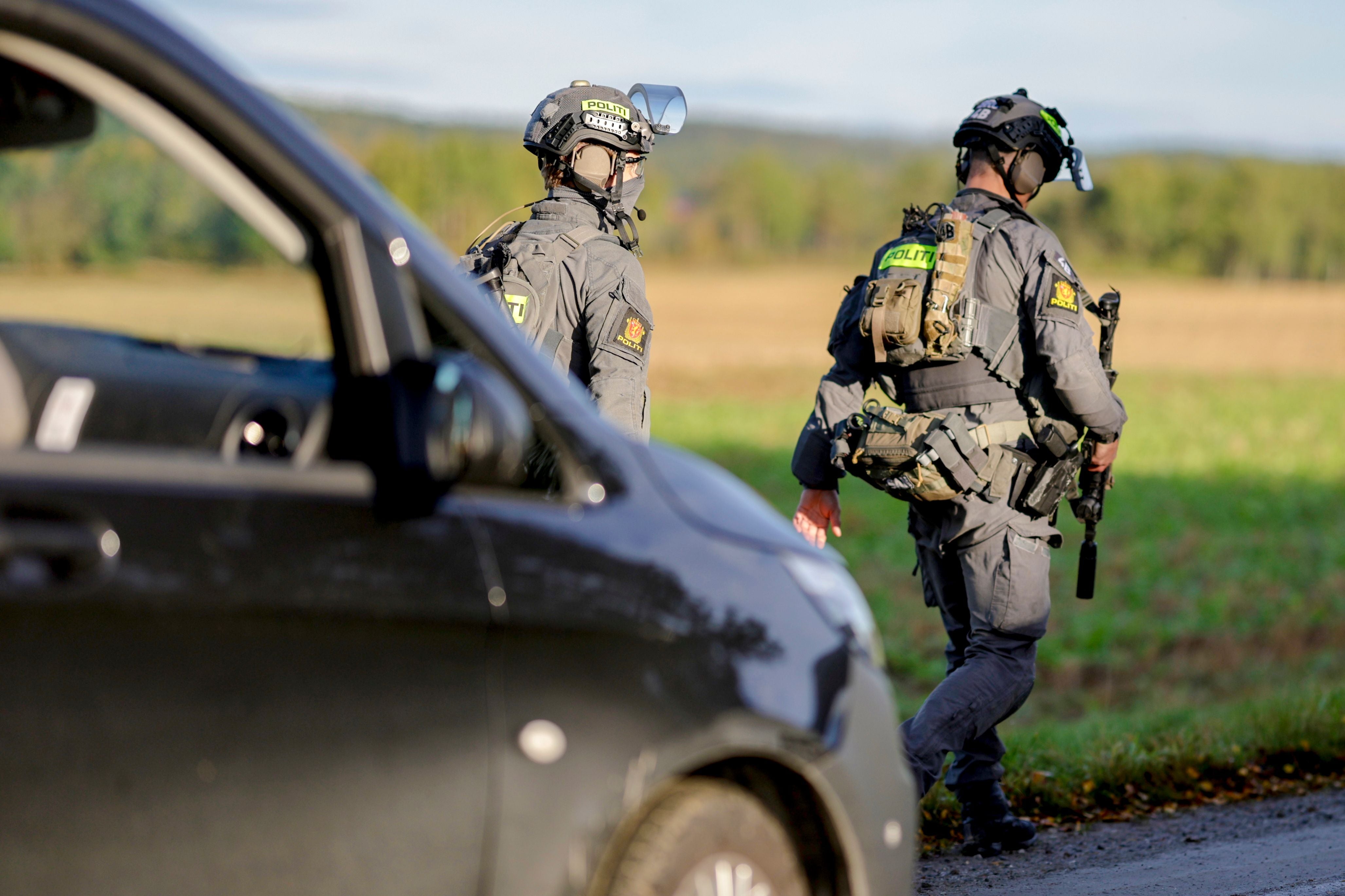 Zwei schwer bewaffnete Polizisten gehen an einem schwarzen Auto auf einer Wiese entlang.