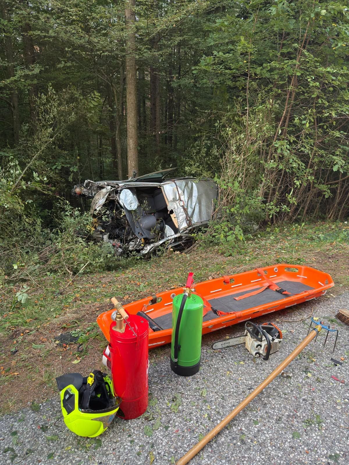 Verunfalltes Auto im Wald, davor Rettungsausrüstung und Trage am Straßenrand.