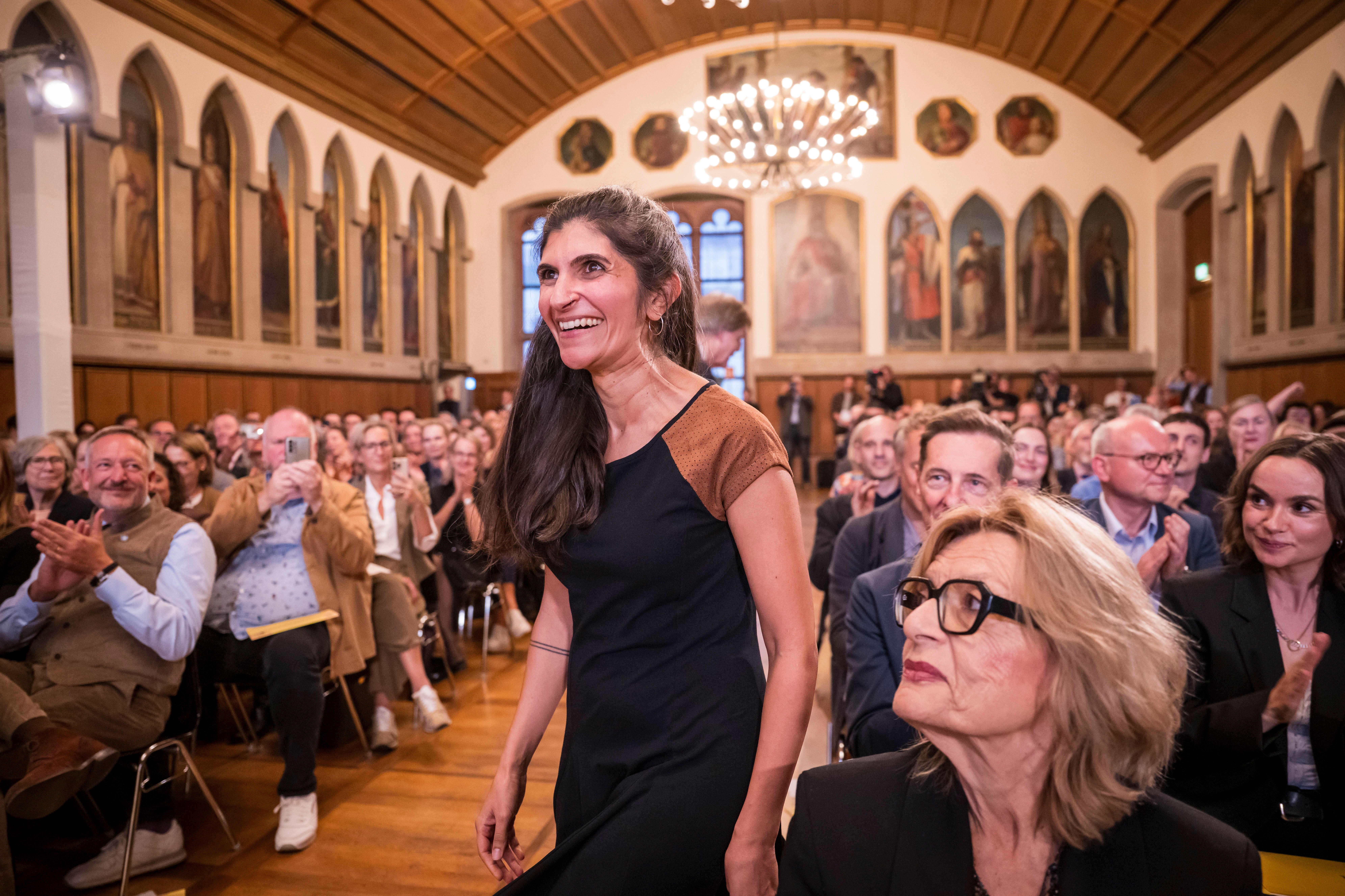 Eine Frau steht lächelnd auf, während das Publikum in einem festlichen Saal applaudiert.