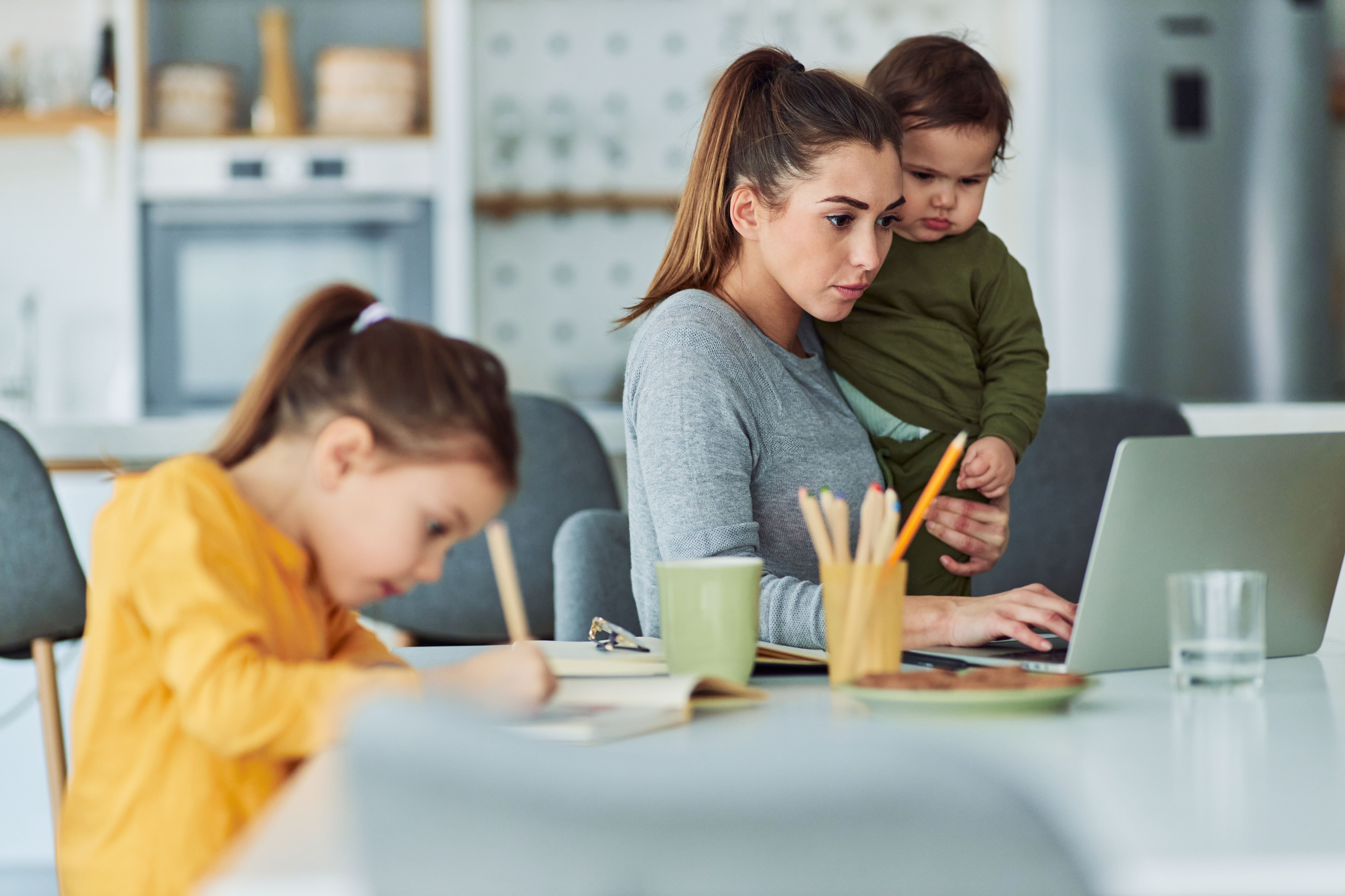 Eine Mutter arbeitet am Laptop, während sie ihr Kleinkind hält und ein weiteres Kind malt.