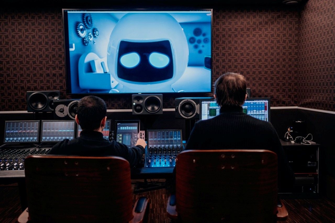 Zwei Personen arbeiten in einem Tonstudio vor Bildschirmen, auf einem Monitor ist ein Roboter zu sehen.