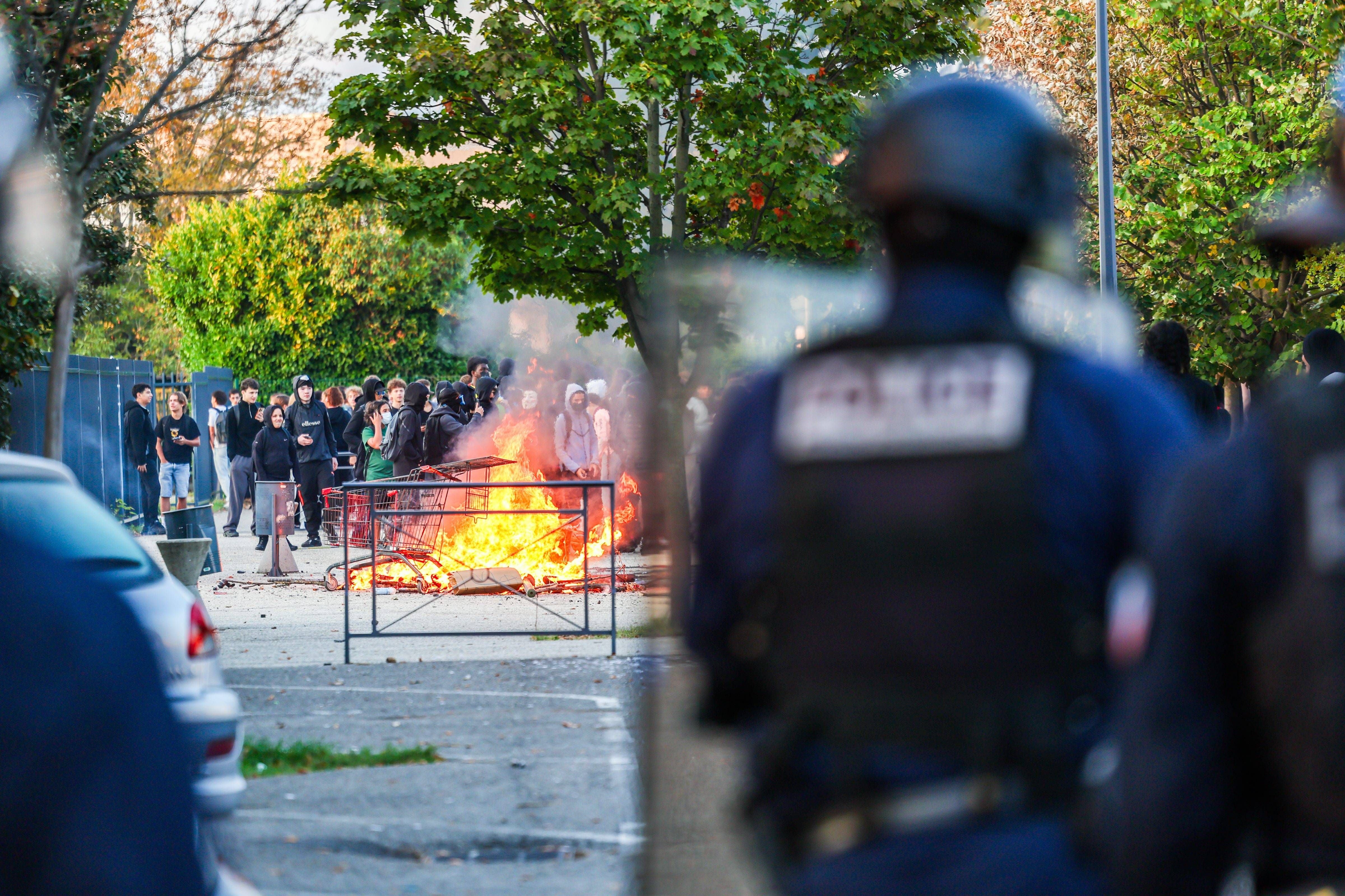 Polizisten stehen vor brennenden Barrikaden, im Hintergrund protestieren zahlreiche Schüler.