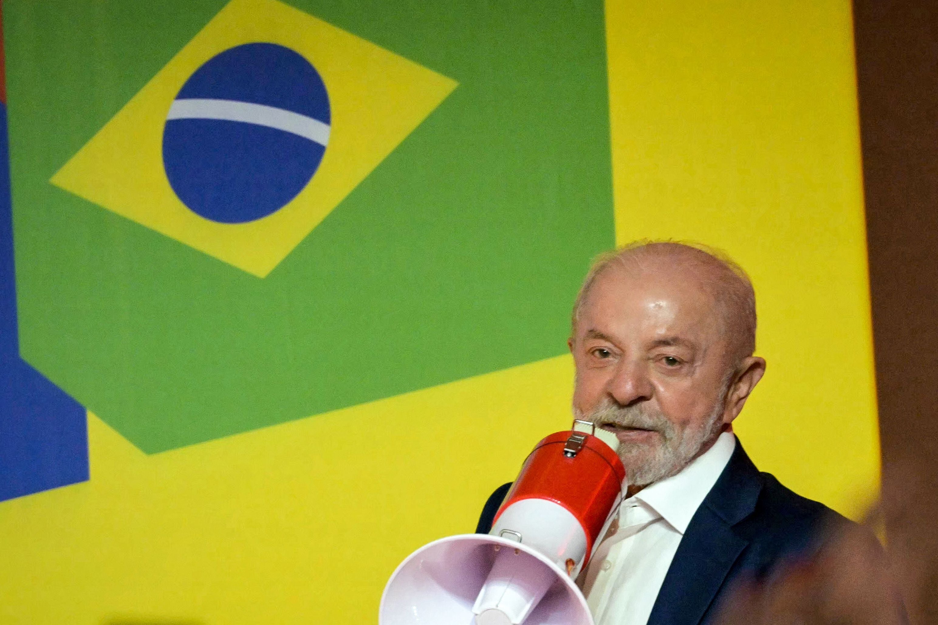 Ein Mann mit Megafon spricht vor einer Wand mit brasilianischer Flagge im Hintergrund.