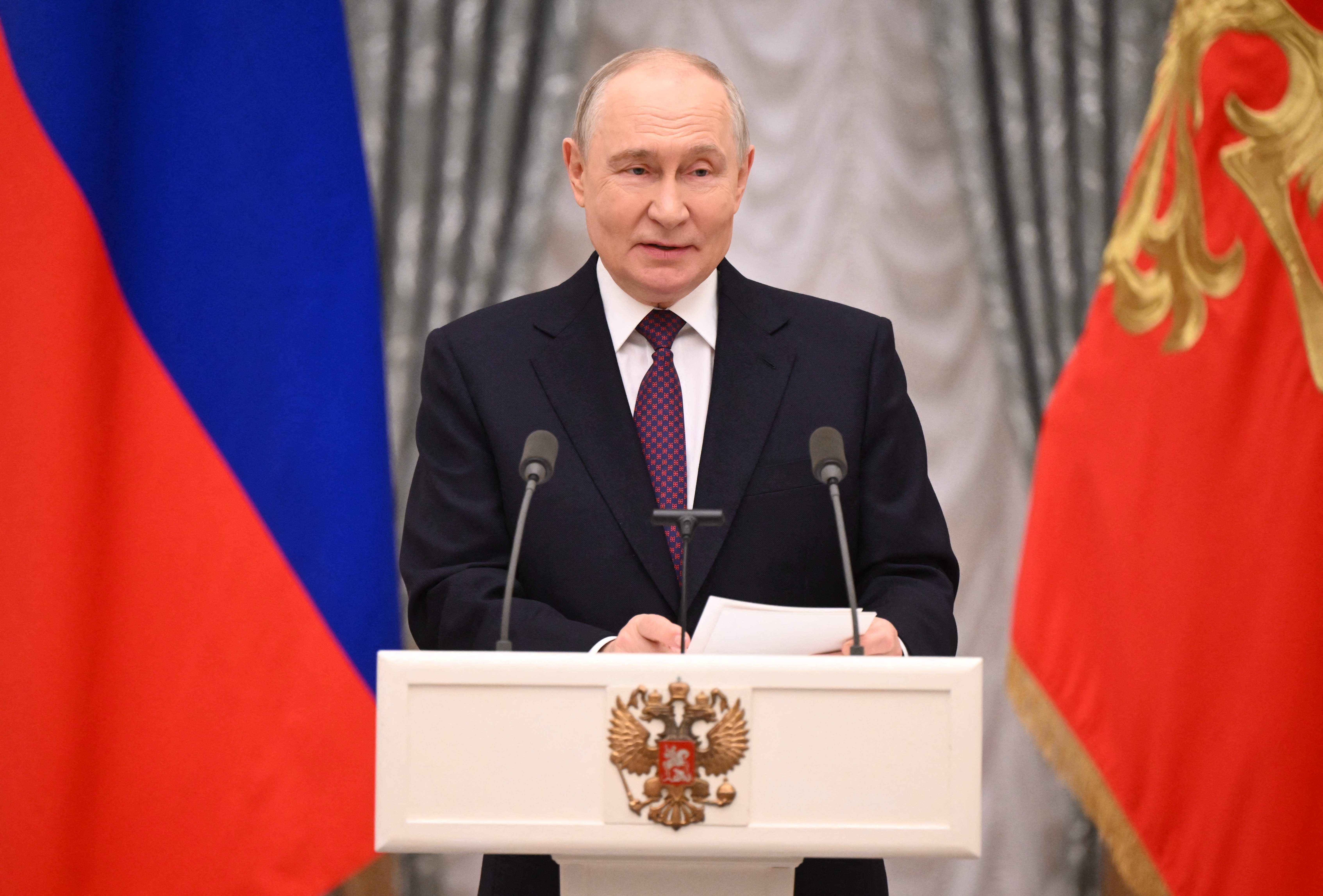Ein Mann im Anzug spricht an einem Rednerpult mit russischem Wappen zwischen zwei Flaggen.