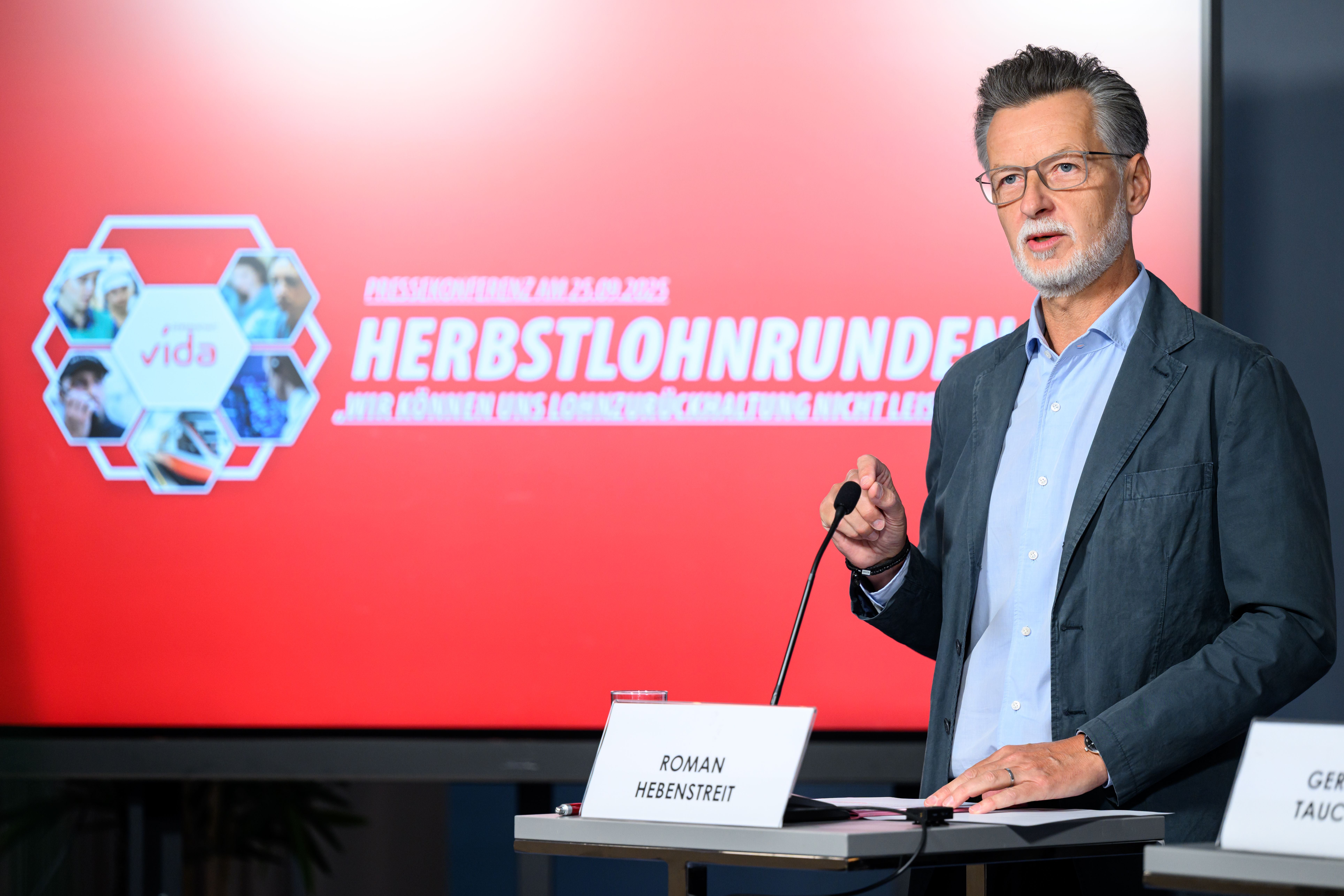 Roman Hebenstreit spricht bei einer Pressekonferenz vor rotem Hintergrund mit "Herbstlohnrunden"-Text.