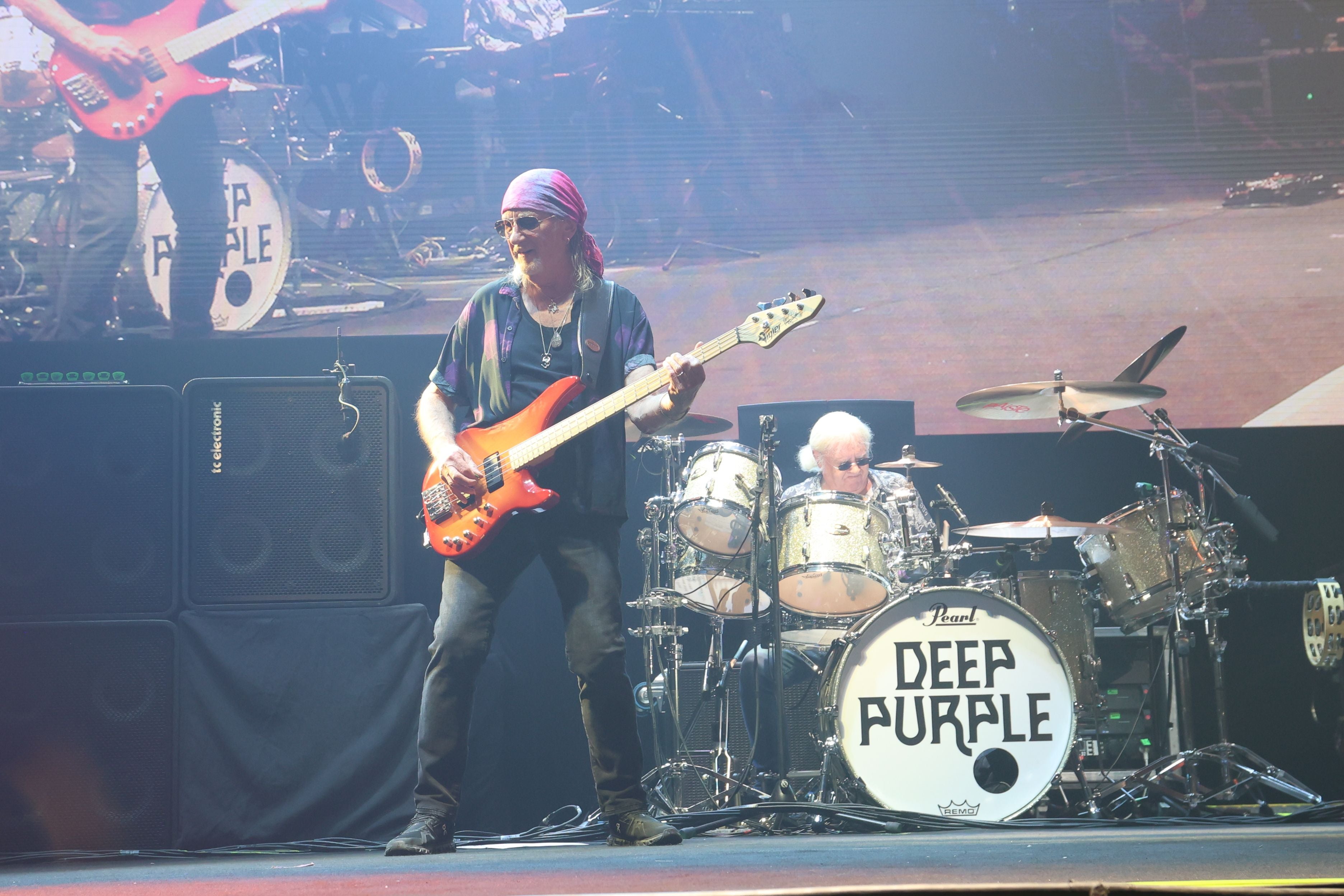 Zwei Deep Purple Musiker spielen Bass und Schlagzeug auf einer Bühne, das Bandlogo ist sichtbar.