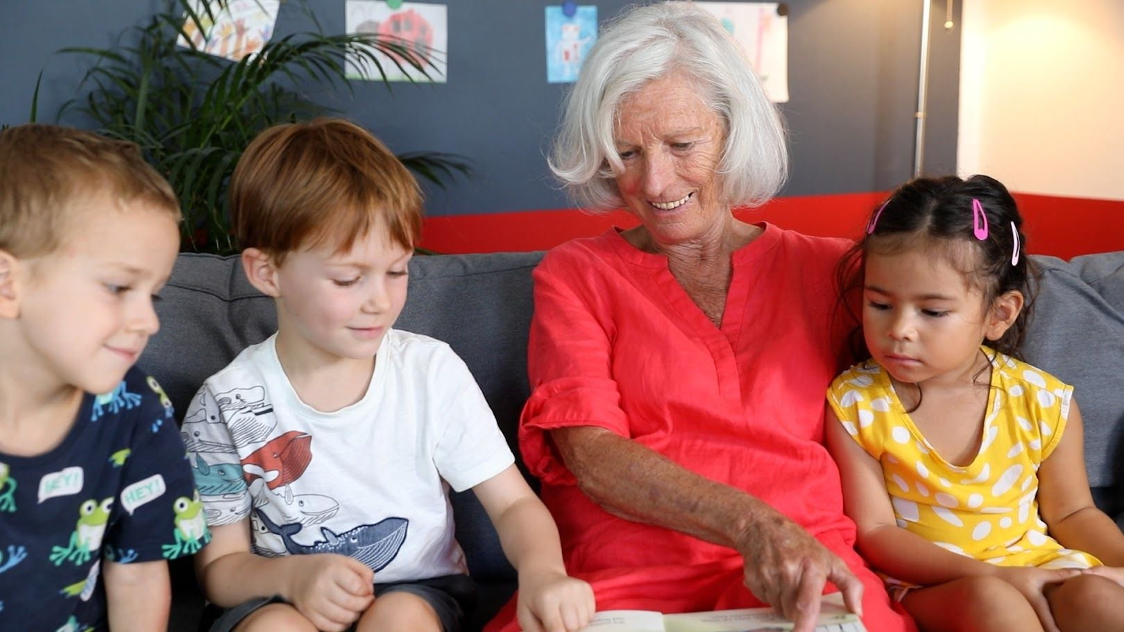 Ältere Frau liest drei Kindern auf einem Sofa gemeinsam aus einem Buch vor.