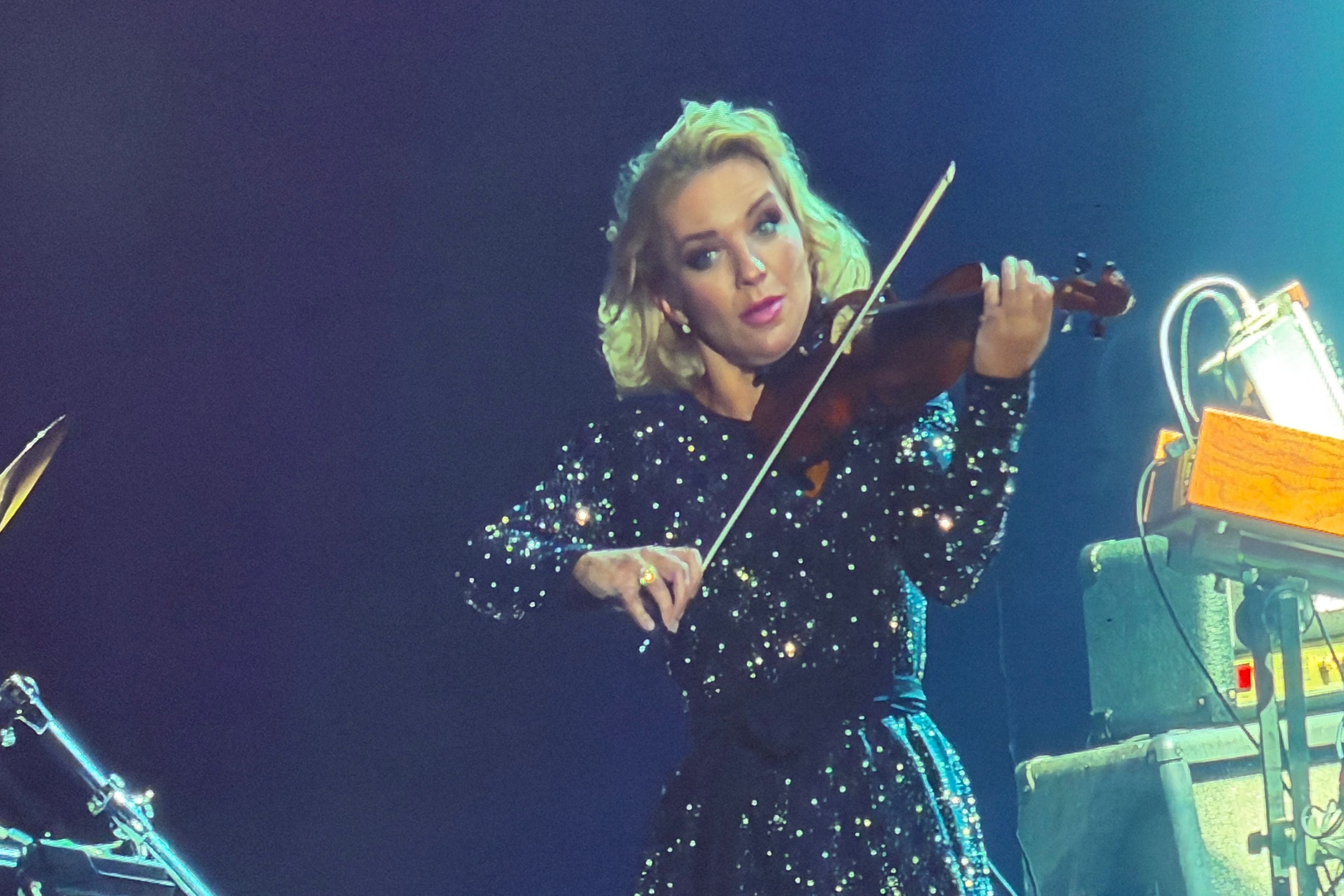 Frau in glitzerndem Kleid spielt eine Violine auf einer beleuchteten Bühne.