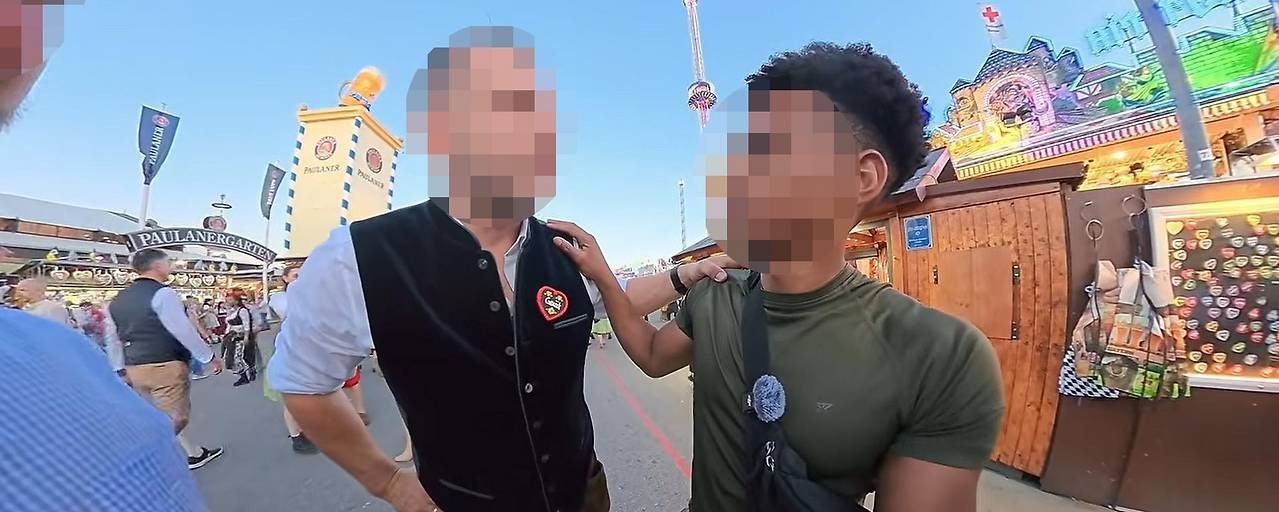 Zwei Männer unterhalten sich auf einem Volksfest mit Ständen und Fahrgeschäften im Hintergrund.