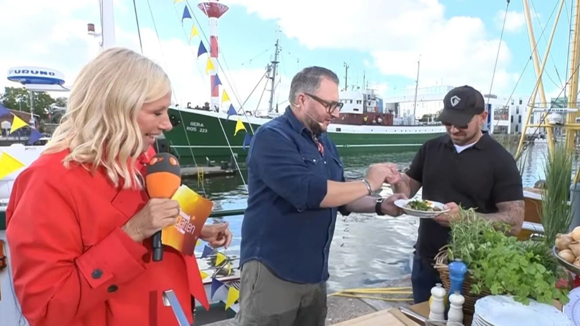 Drei Personen stehen am Hafen, eine moderiert, zwei probieren Essen, im Hintergrund Schiffe.