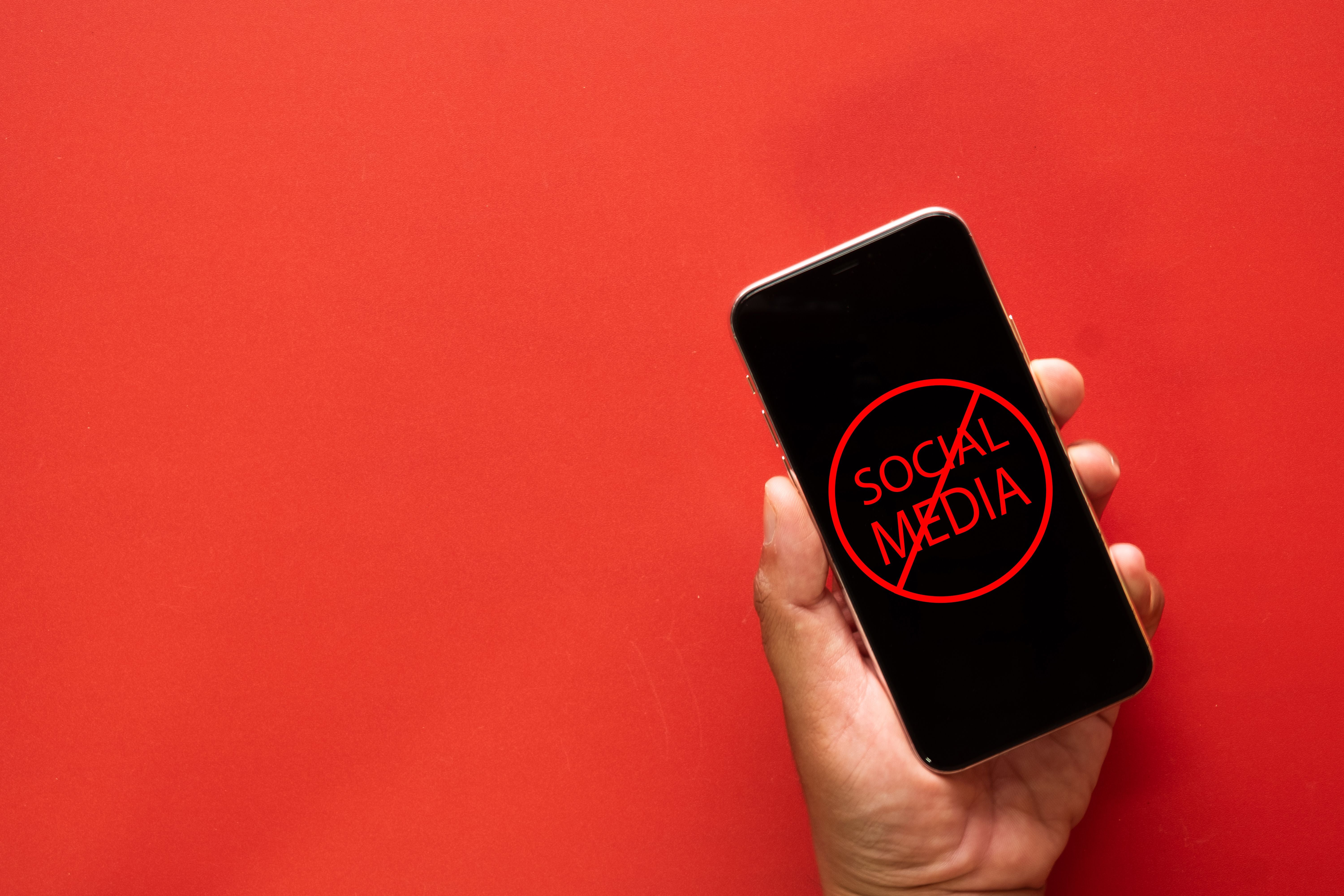 Hand hält Smartphone mit durchgestrichenem „Social Media“ auf rotem Hintergrund.