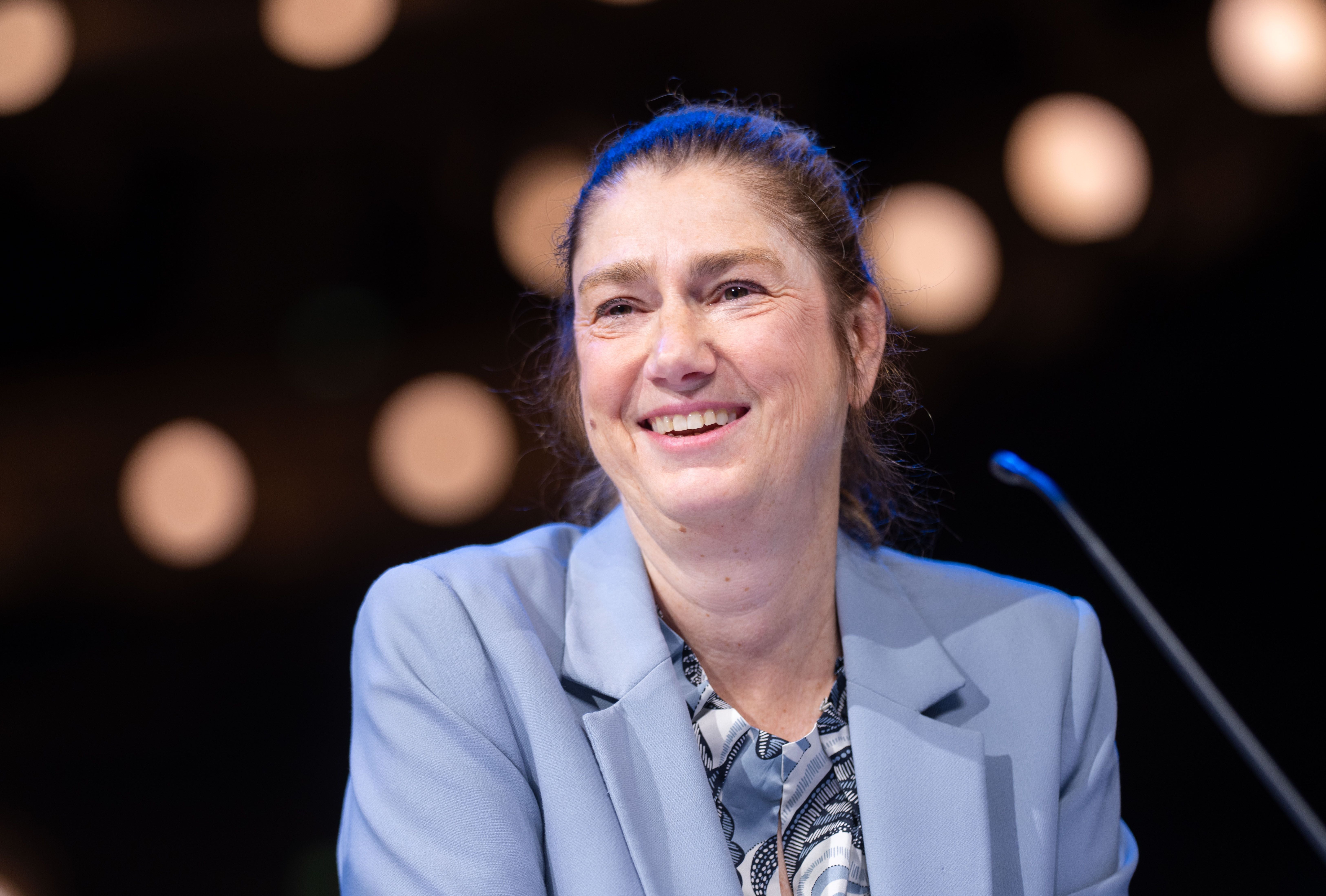 Frau mit hellblauem Blazer lächelt auf einer Bühne vor unscharfem Hintergrund mit Lichtern.