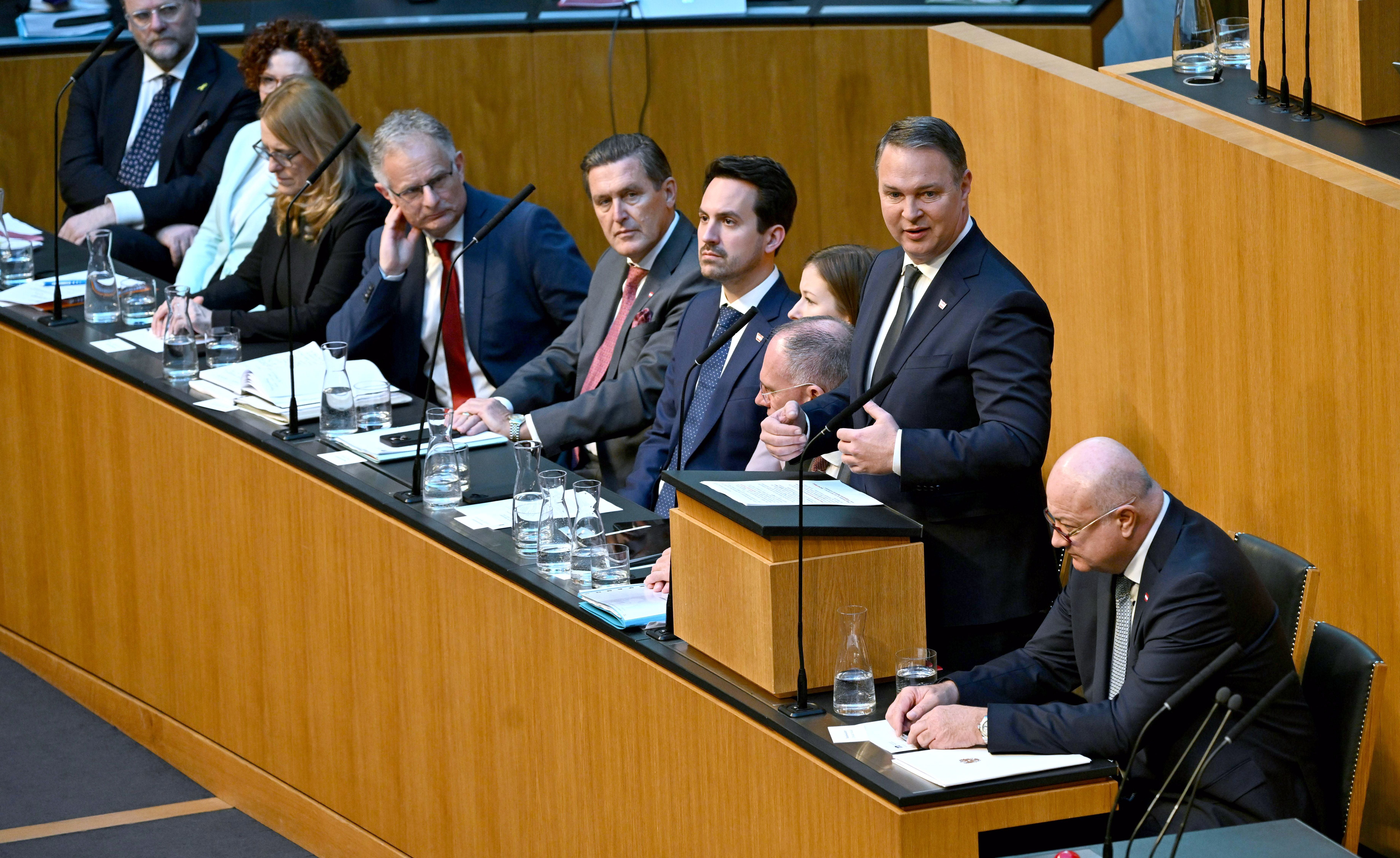 Mehrere Politiker sitzen und stehen an der Regierungsbank im österreichischen Parlament bei einer Sitzung.