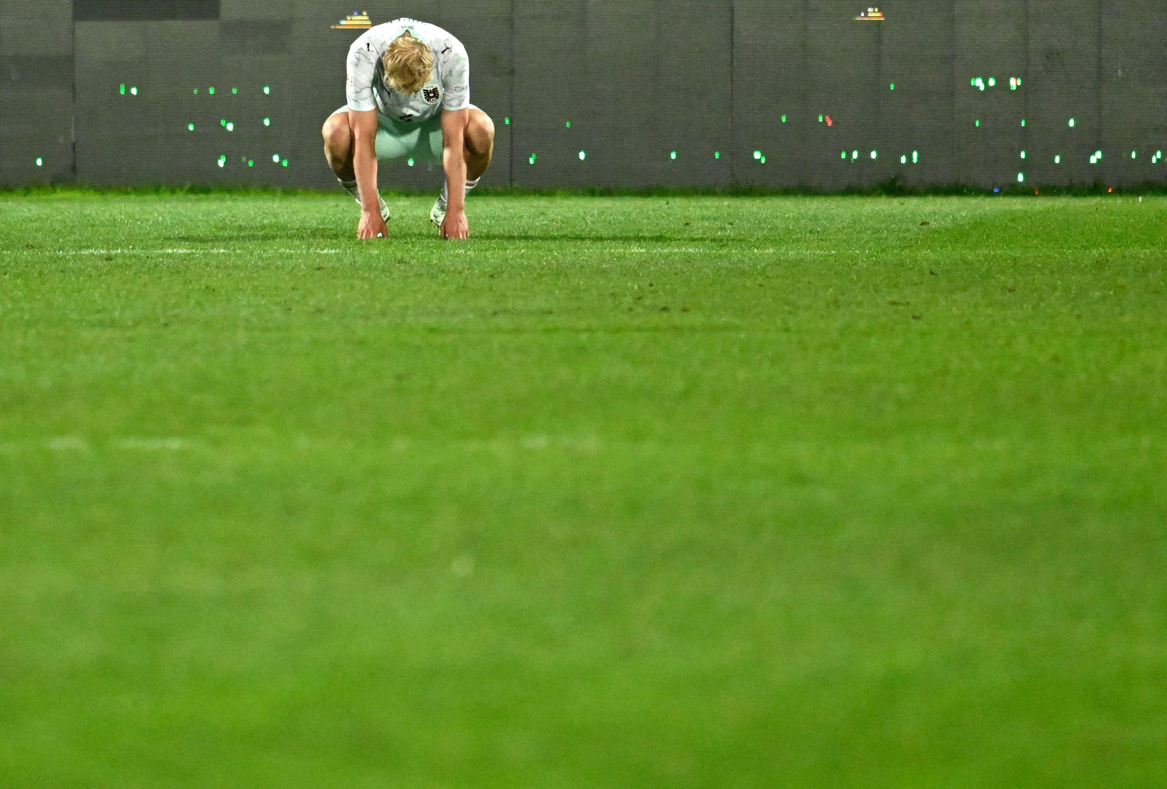 Ein Fußballspieler hockt enttäuscht auf dem Spielfeld, viel leere Rasenfläche im Vordergrund.