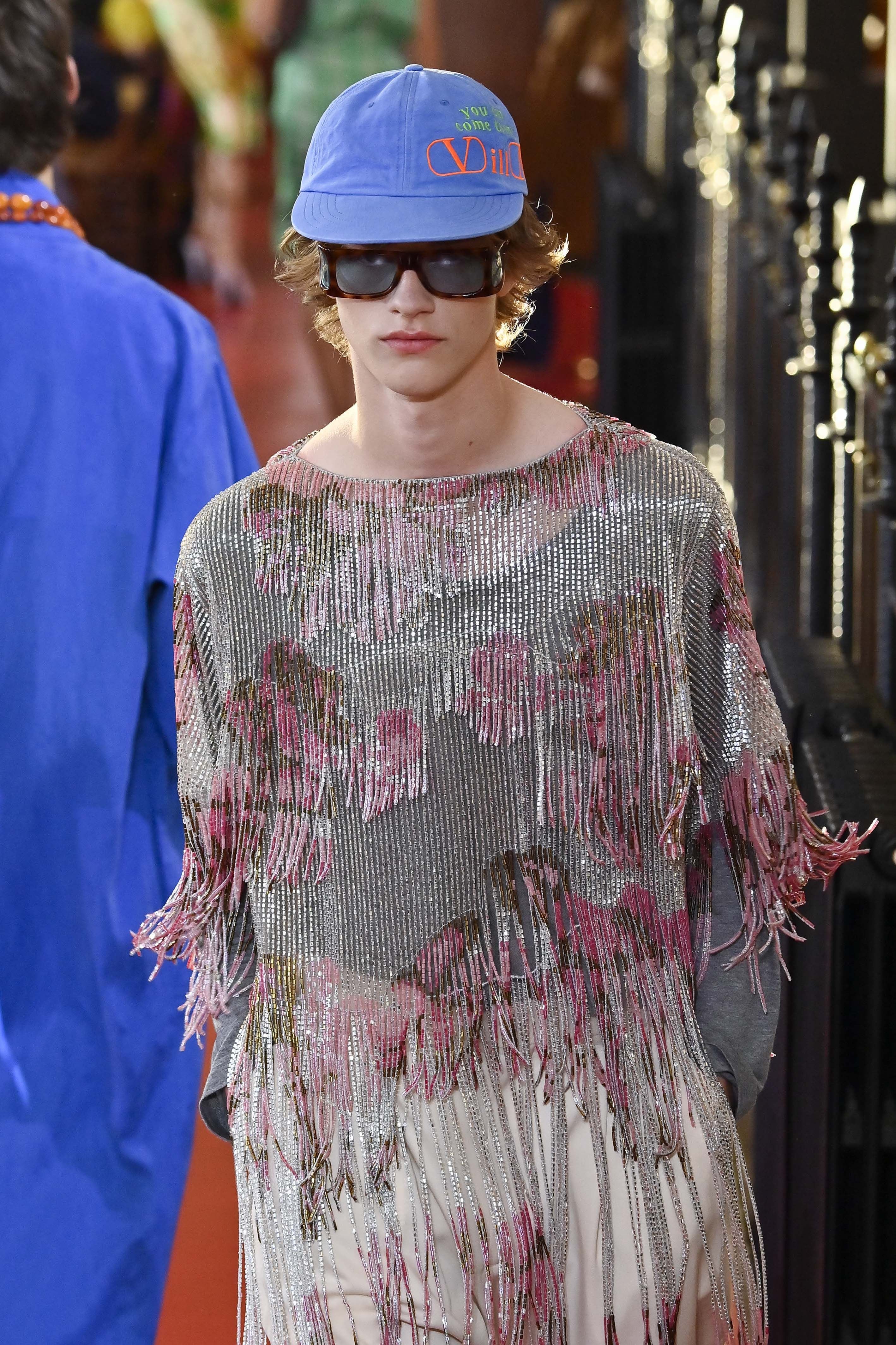 Model mit blauem Cap, Sonnenbrille und silbernem Fransentop auf einer Modenschau-Laufsteg.