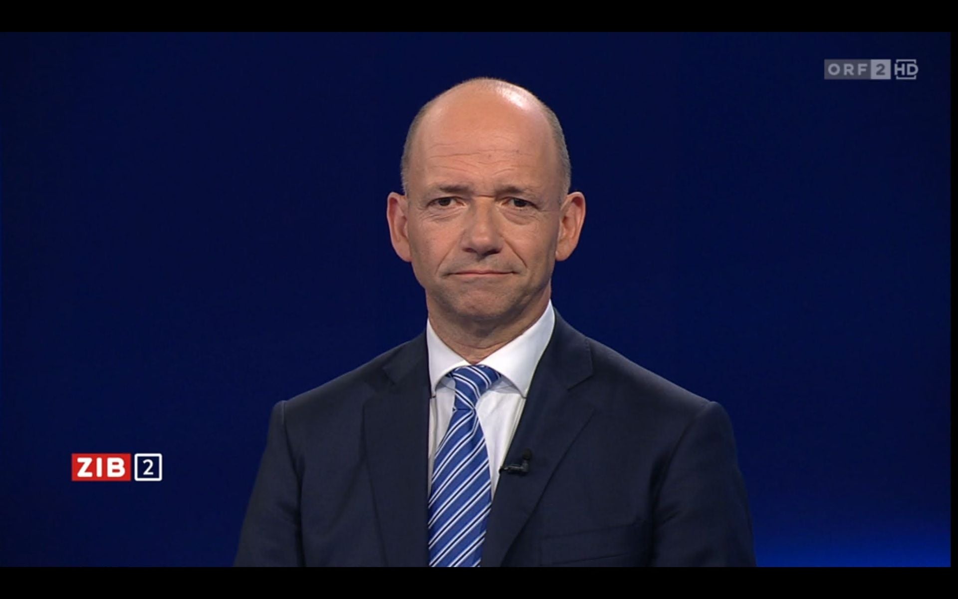 Ein Mann im Anzug mit gestreifter Krawatte steht vor blauem Hintergrund im Studio.